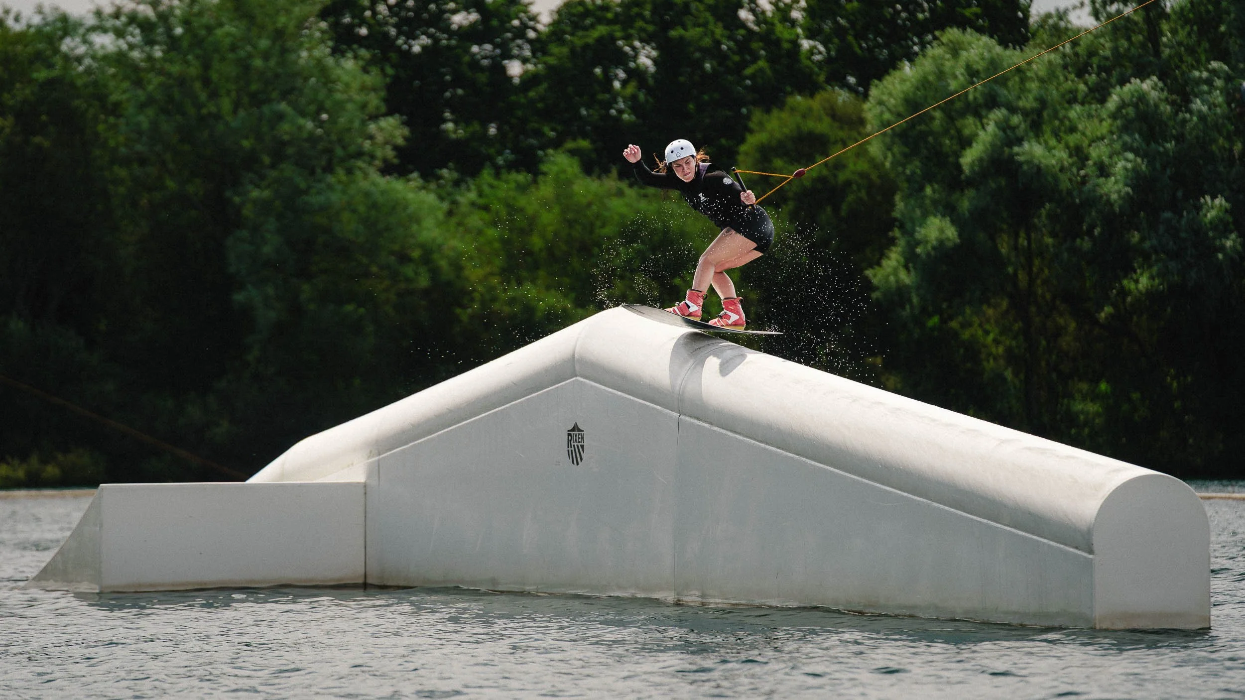 A woman in a helmet and black wetsuit wakeboarding on a water ramp, with trees in the background.