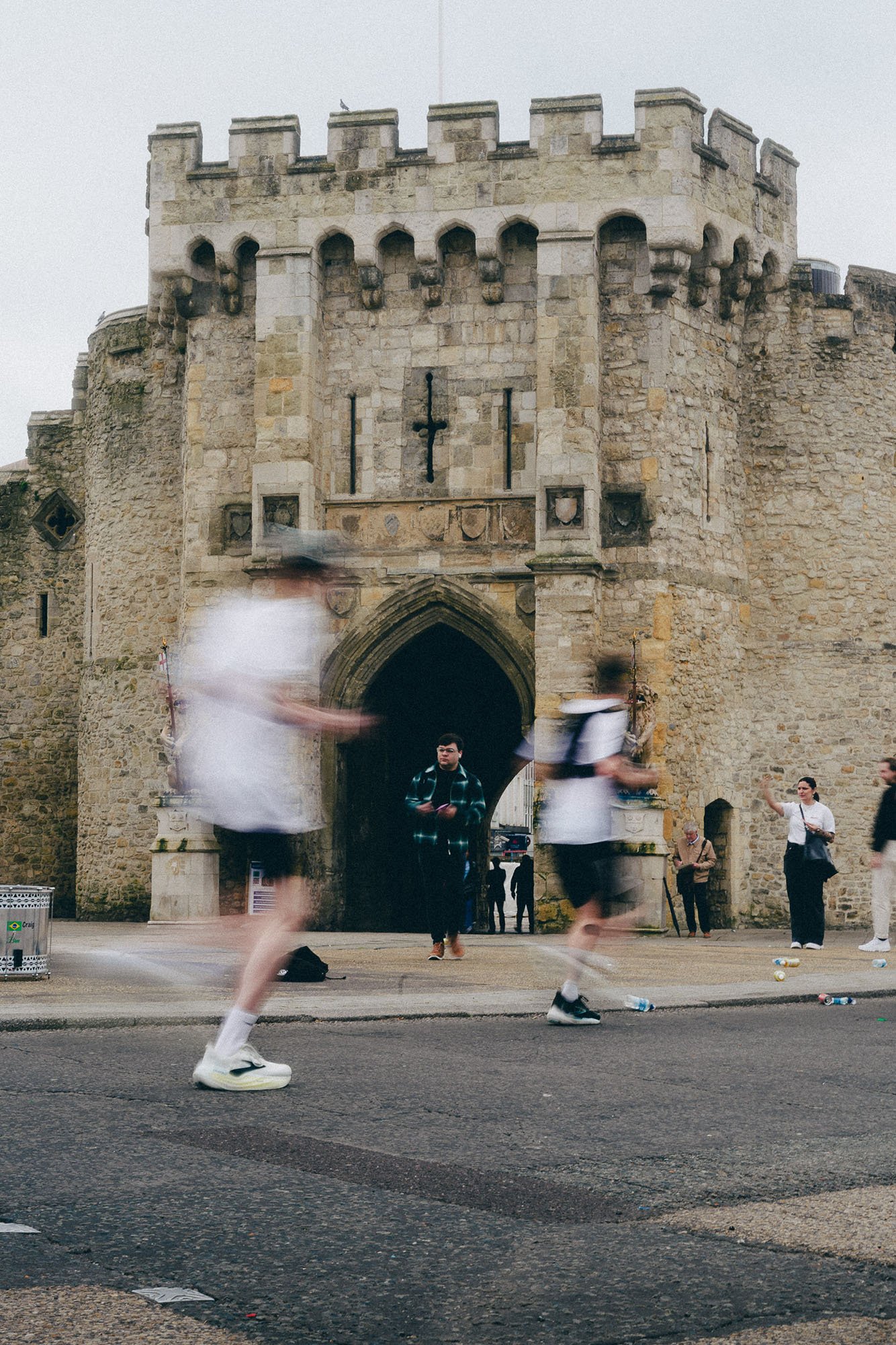 runners blurred as they run past an old stone castle gate with a cross above the archway, some standing and some walking, in a city street.