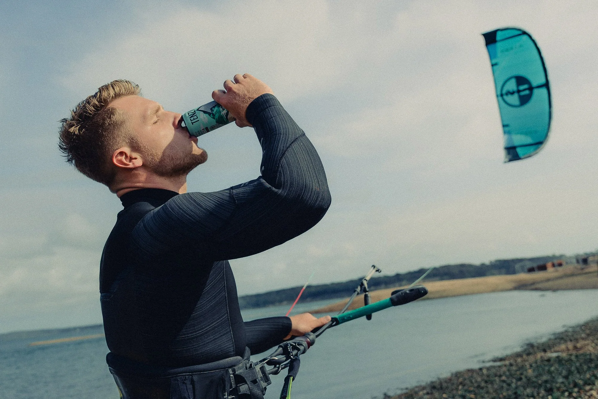 A kitesurfer with short, blonde hair, wearing a black wetsuit, is standing near a body of water, holding a kiteboarding control bar with two lines, with a kite in the distance. He is drinking from a Tenzing natural energy drink can.