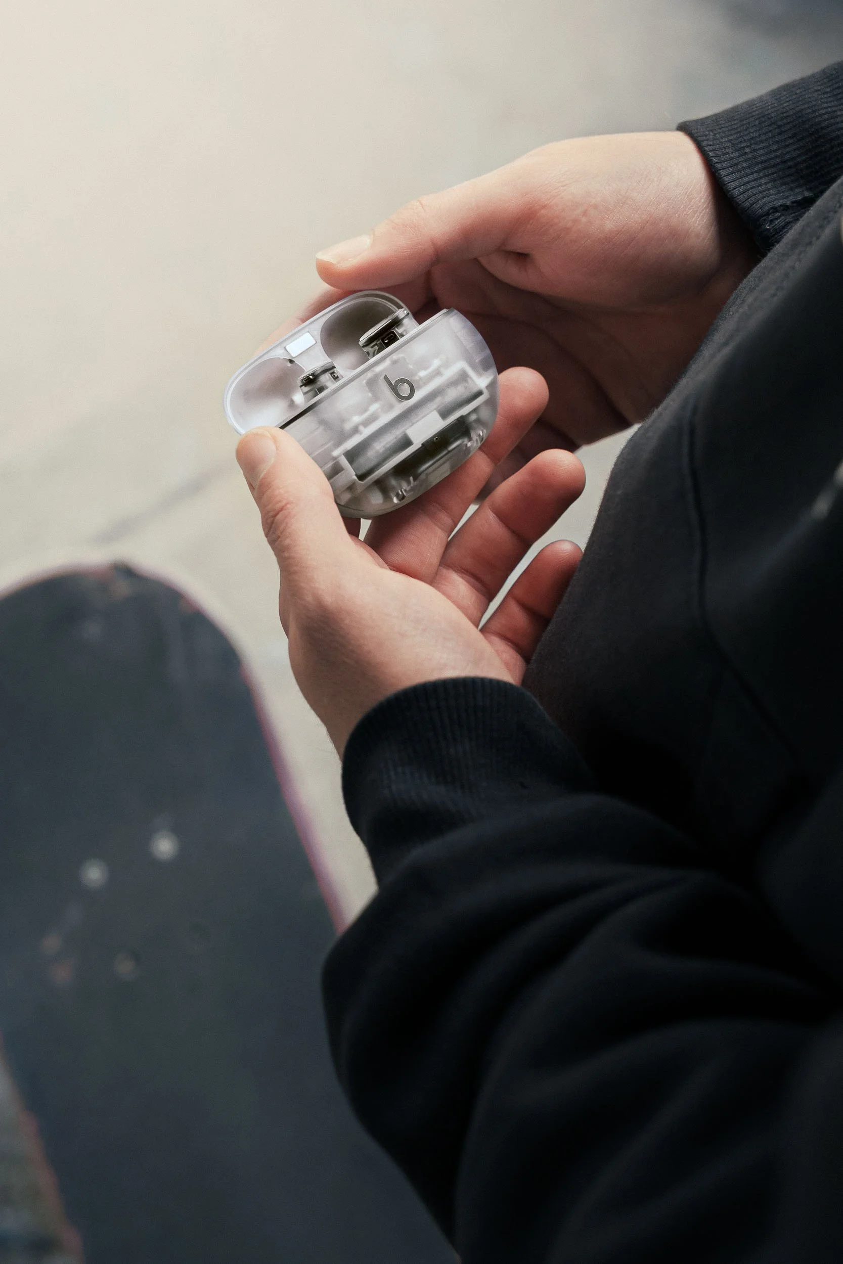 Close-up of a person holding a wireless earbuds case, with earbuds inside, in a person's hands. The person is wearing a black long-sleeved hoodie.