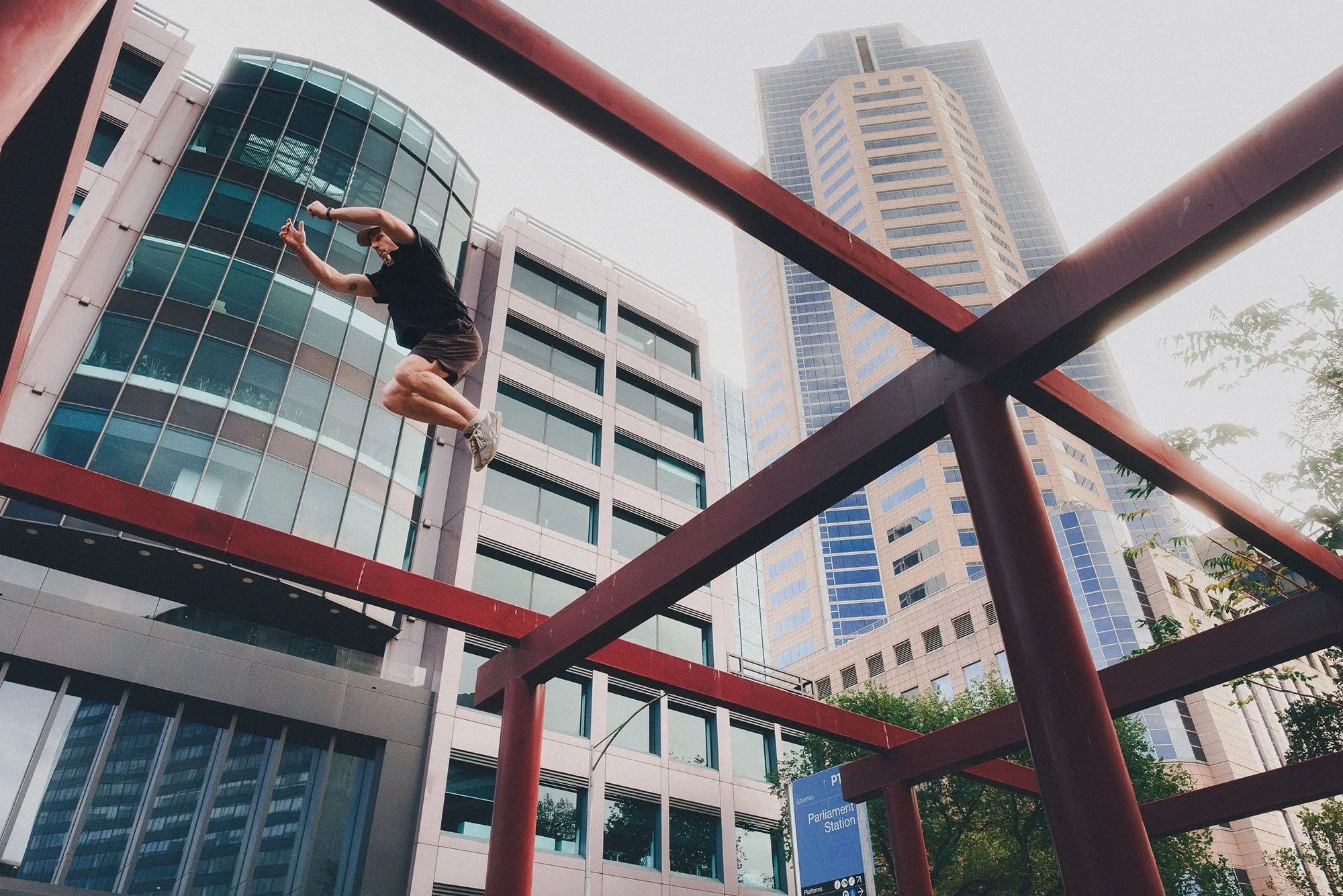 A person in black clothing jumping between red steel beams with tall modern buildings in the background in an urban area.
