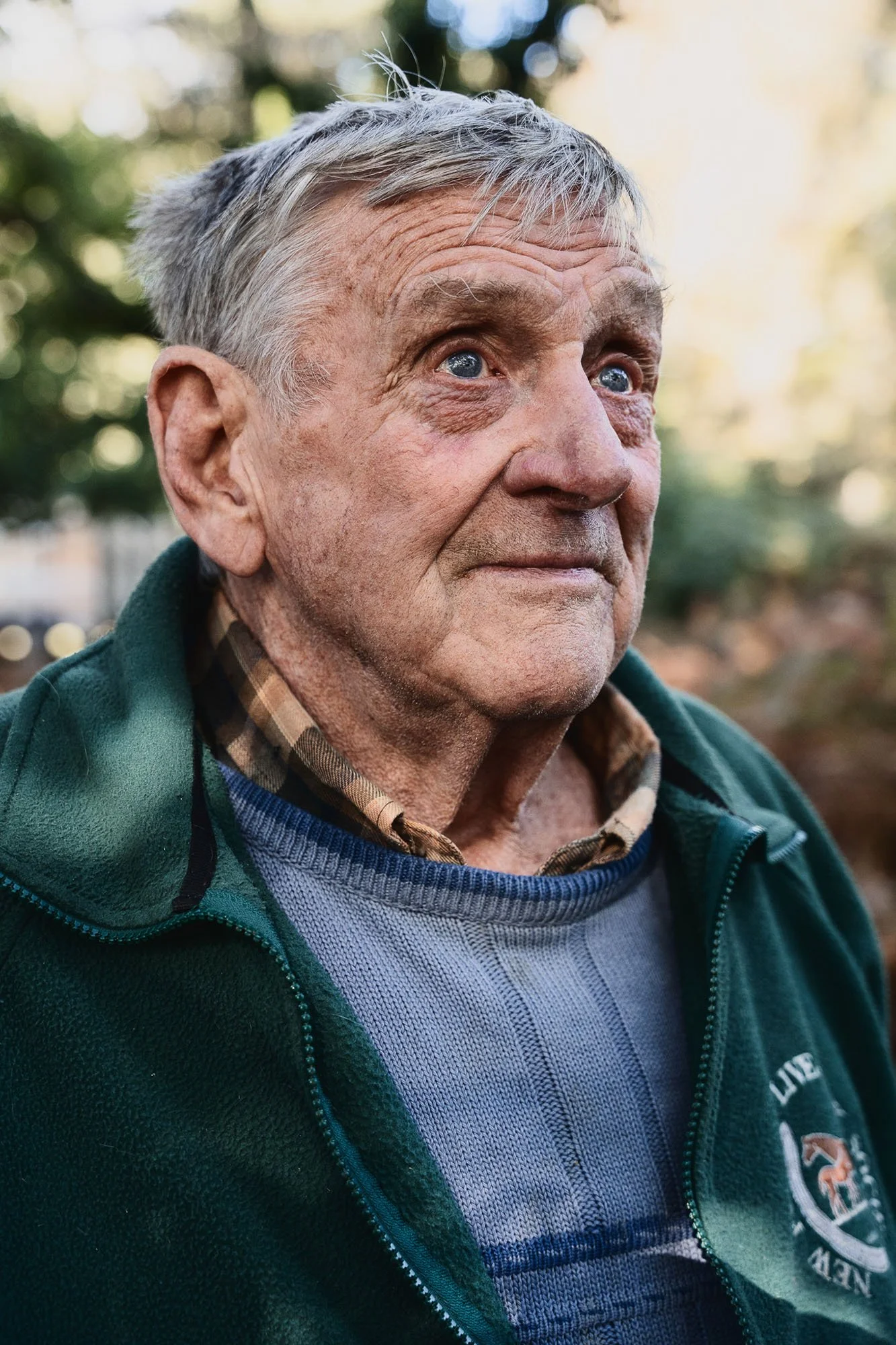 An elderly man with gray hair and blue eyes is outdoors, wearing a green jacket over a plaid shirt and a blue sweater. The background is blurred with trees and sunlight.