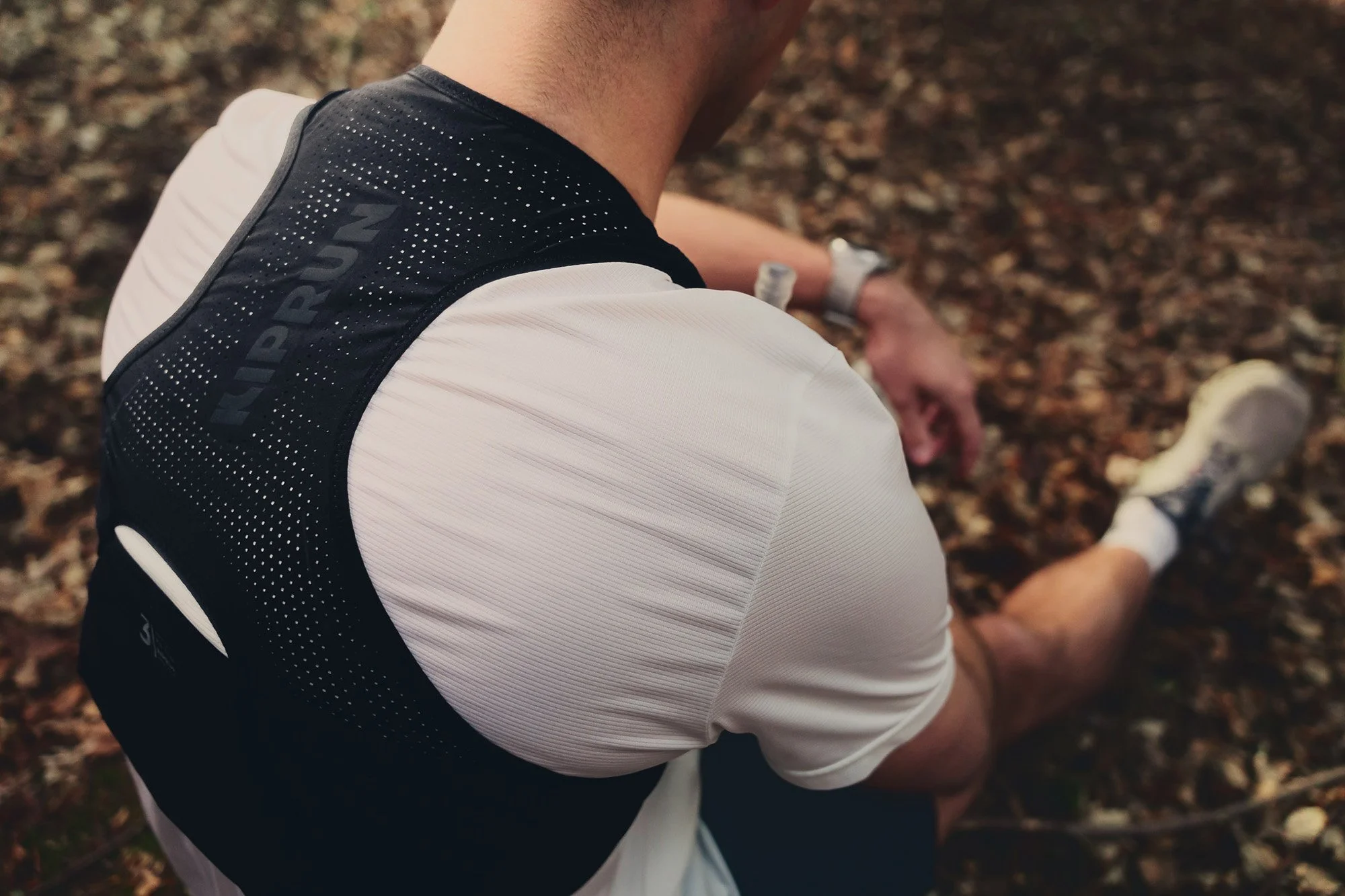 A runner wearing a black Kiprun running vest with the logo 'KIPRUN' vertically on the back, sitting outdoors on a ground covered with leaves, with his hand resting on his knee.
