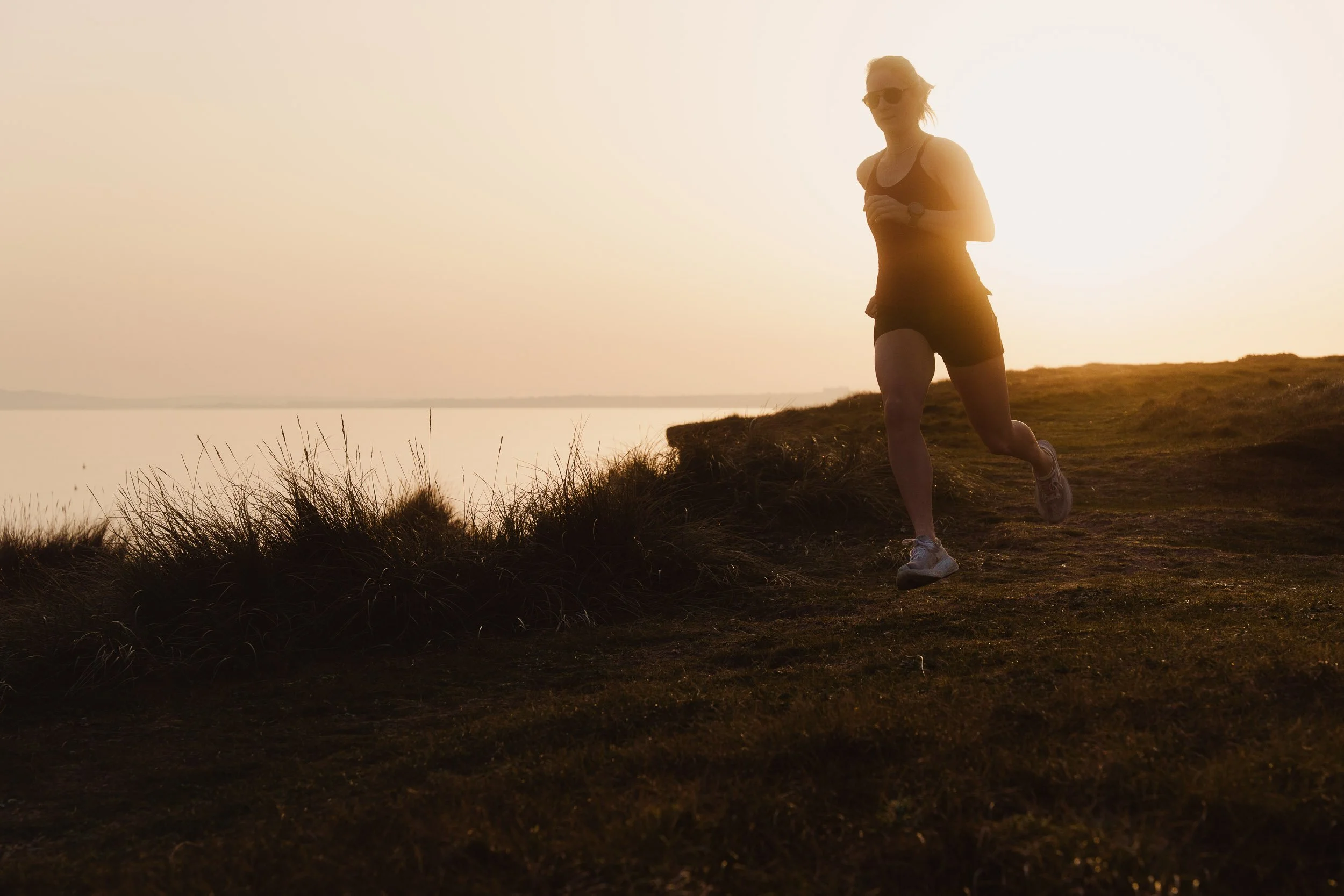 A female runner jogging on a trail near water during sunset, wearing sunglasses, a tank top, shorts, and running shoes.