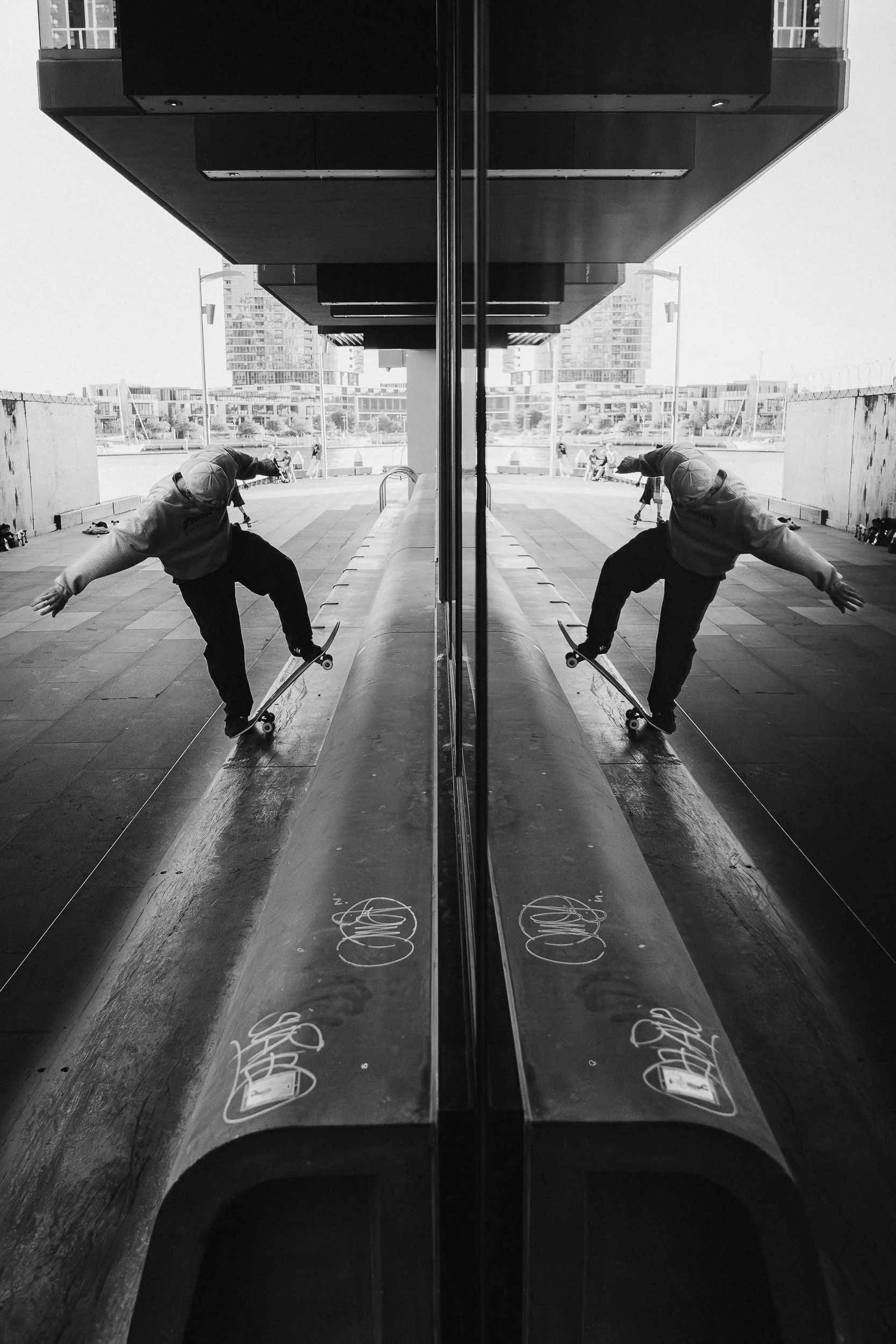 Black and white photo of a city street with a reflective glass building and a person skateboarding on the sidewalk in front of it.