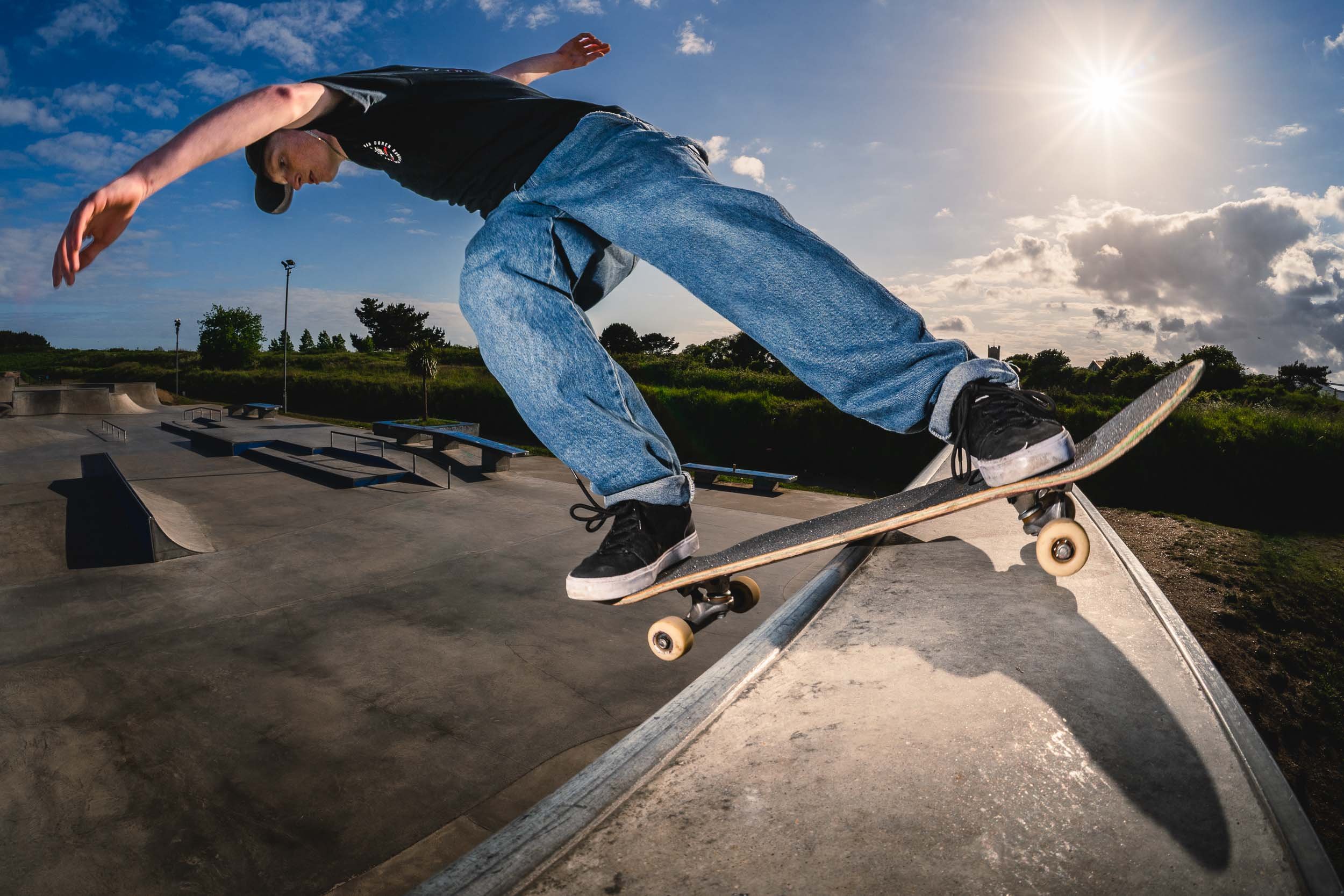Skateboarder performing a trick at a skatepark during sunset, with a partly cloudy sky and green trees in the background.