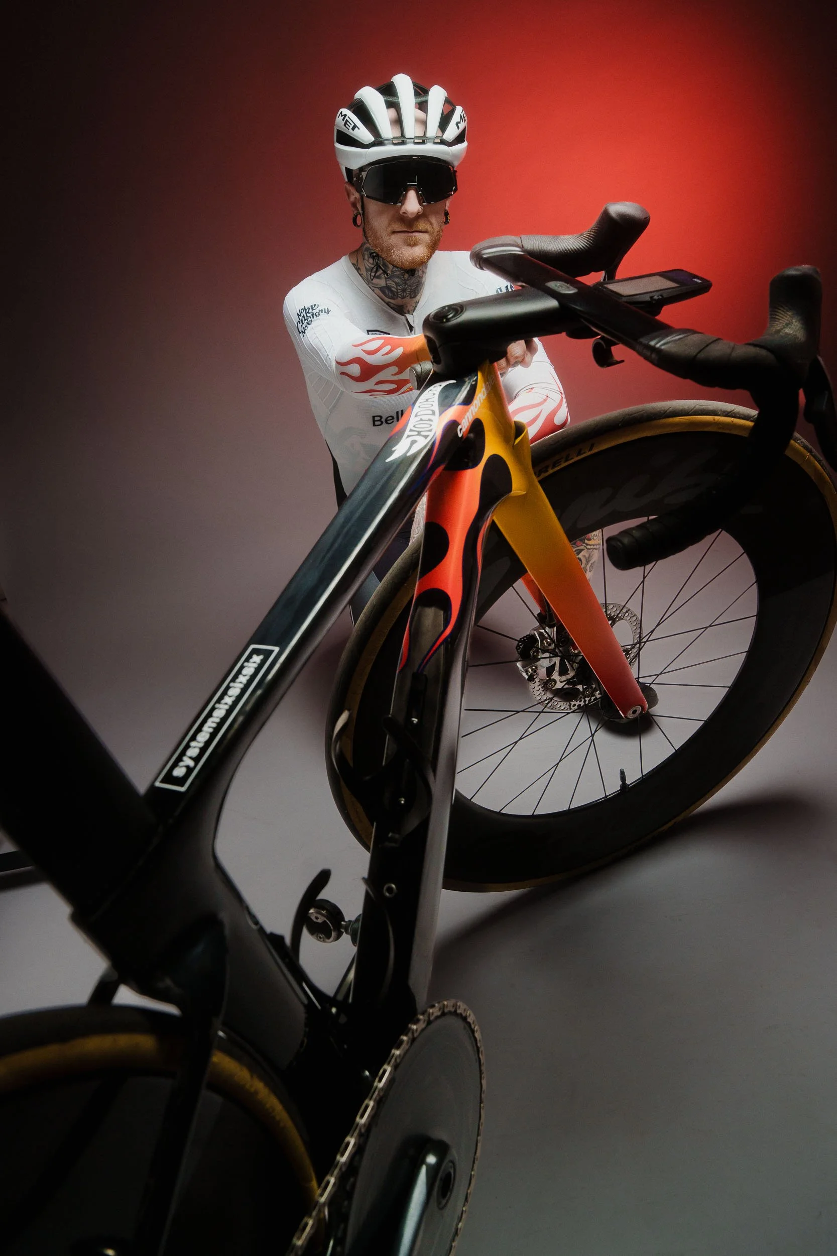 A person wearing a white cycling jersey with flames, a white helmet, and sunglasses, standing next to a black road bicycle with a flame design on it, against a red and grey gradient background.