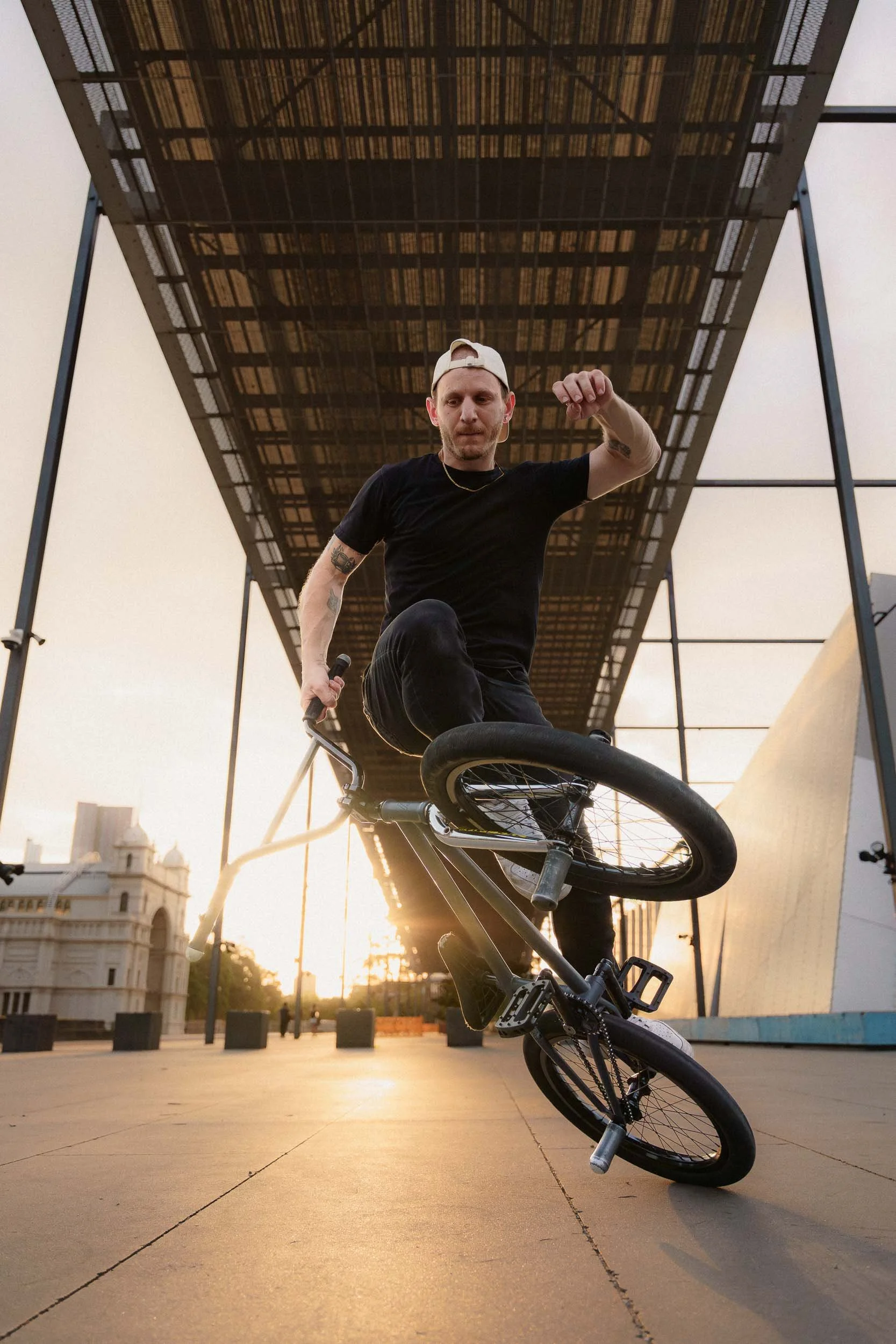 A BMX rider doing flatland BMX tricks under a modern architecture at sunset.