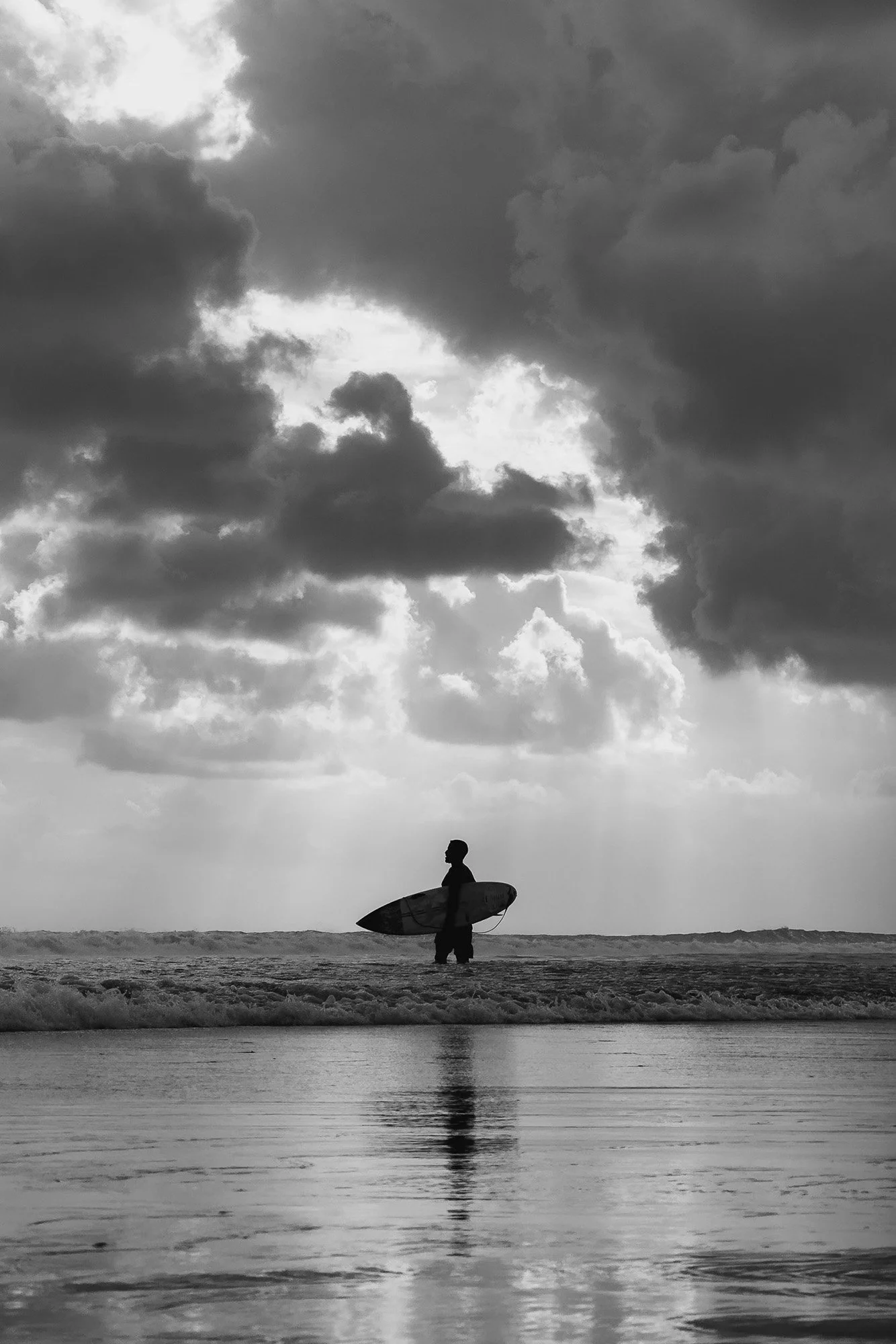 A black and white photo of a person standing on the beach holding a surfboard, looking at the ocean with cloudy skies overhead.