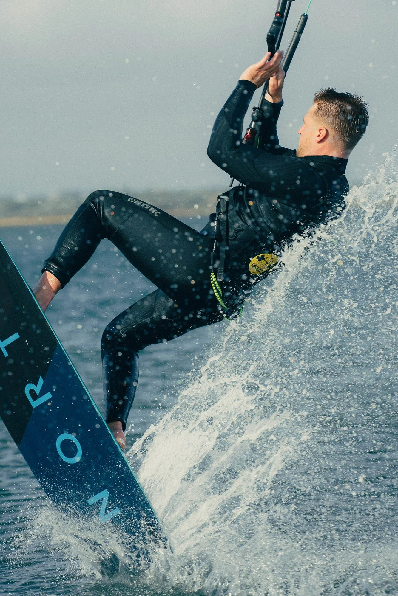A kitesurfer, doing tricks holding onto the control bar with both hands, wearing a wetsuit, with water splashing around him 