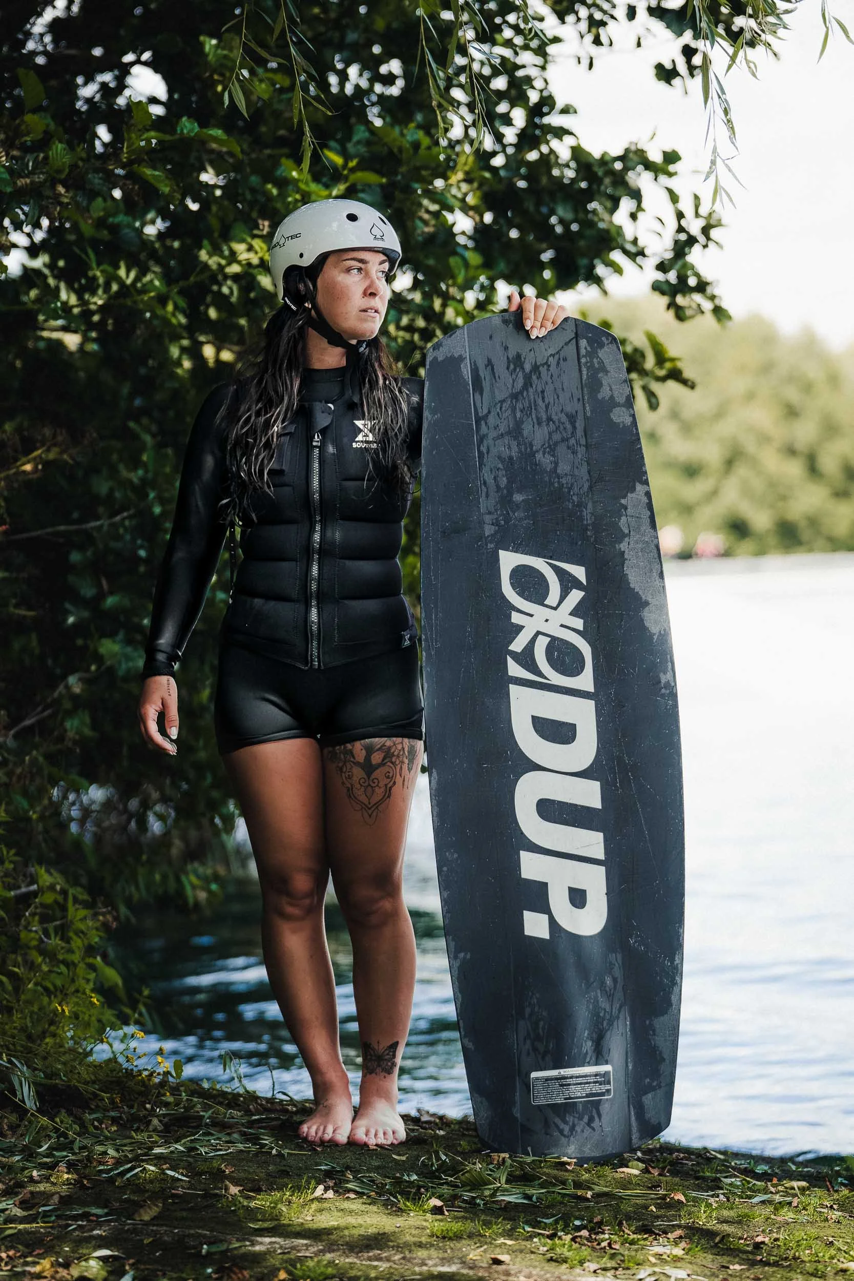 A woman with long dark hair, tattoos on her legs, and a serious expression, wearing a helmet and wetsuit, stands beside a wakeboard on the edge of a body of water surrounded by trees and foliage.