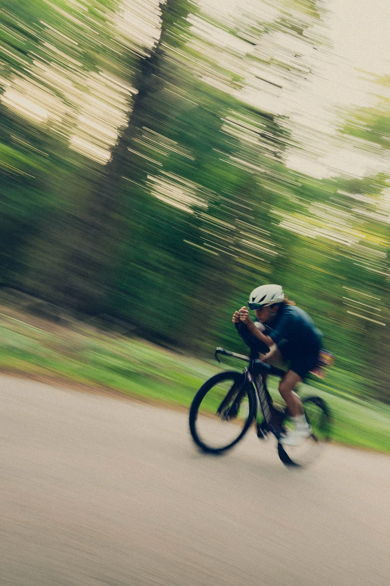 A person riding a bicycle on a road, wearing a white helmet and navy clothing, with motion blur creating a sense of speed and movement, set against a background of trees and greenery.