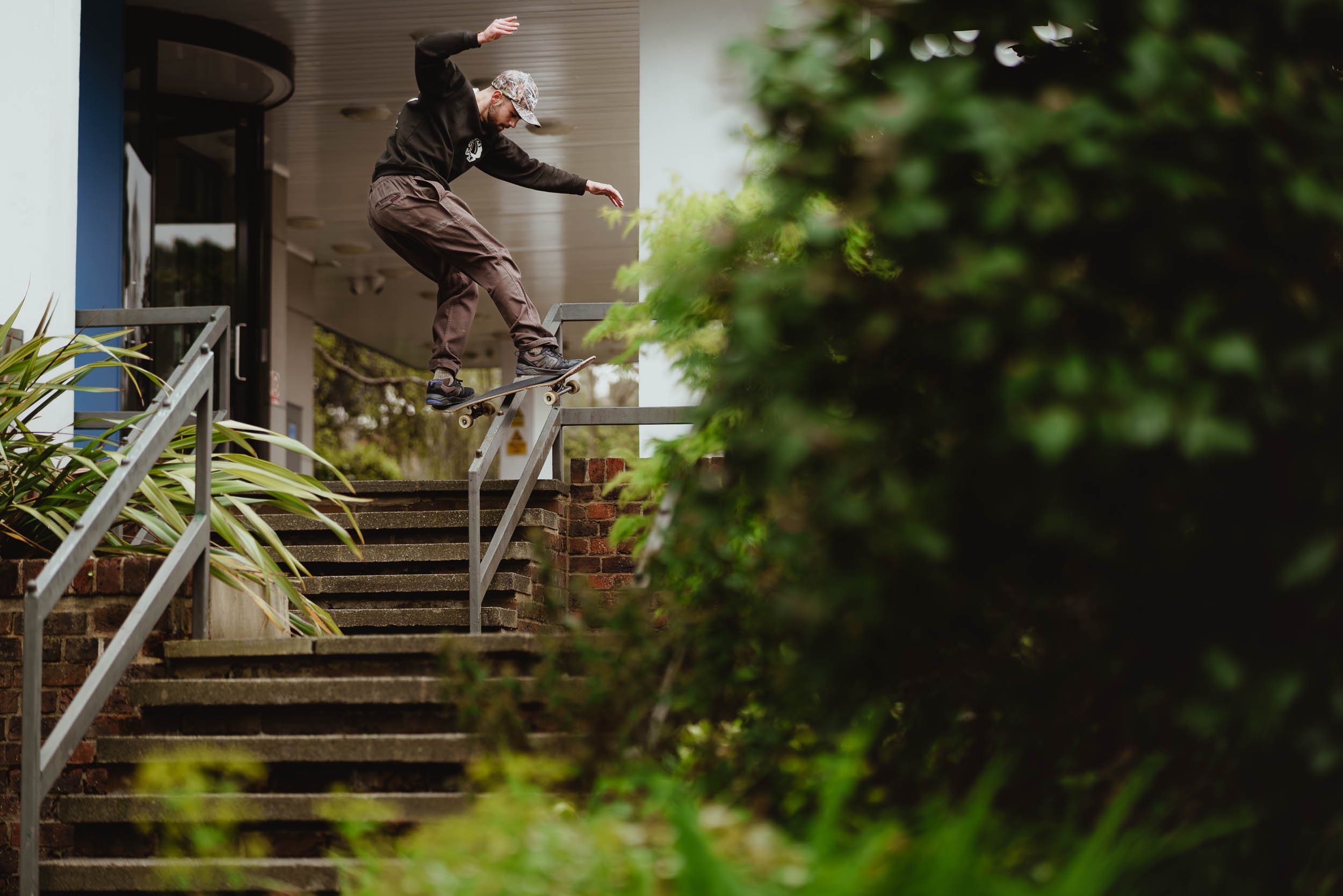 A skateboarder wearing a black hoodie, brown pants, and a camouflage cap performing a trick on outdoor stairs with metal handrails, surrounded by greenery.