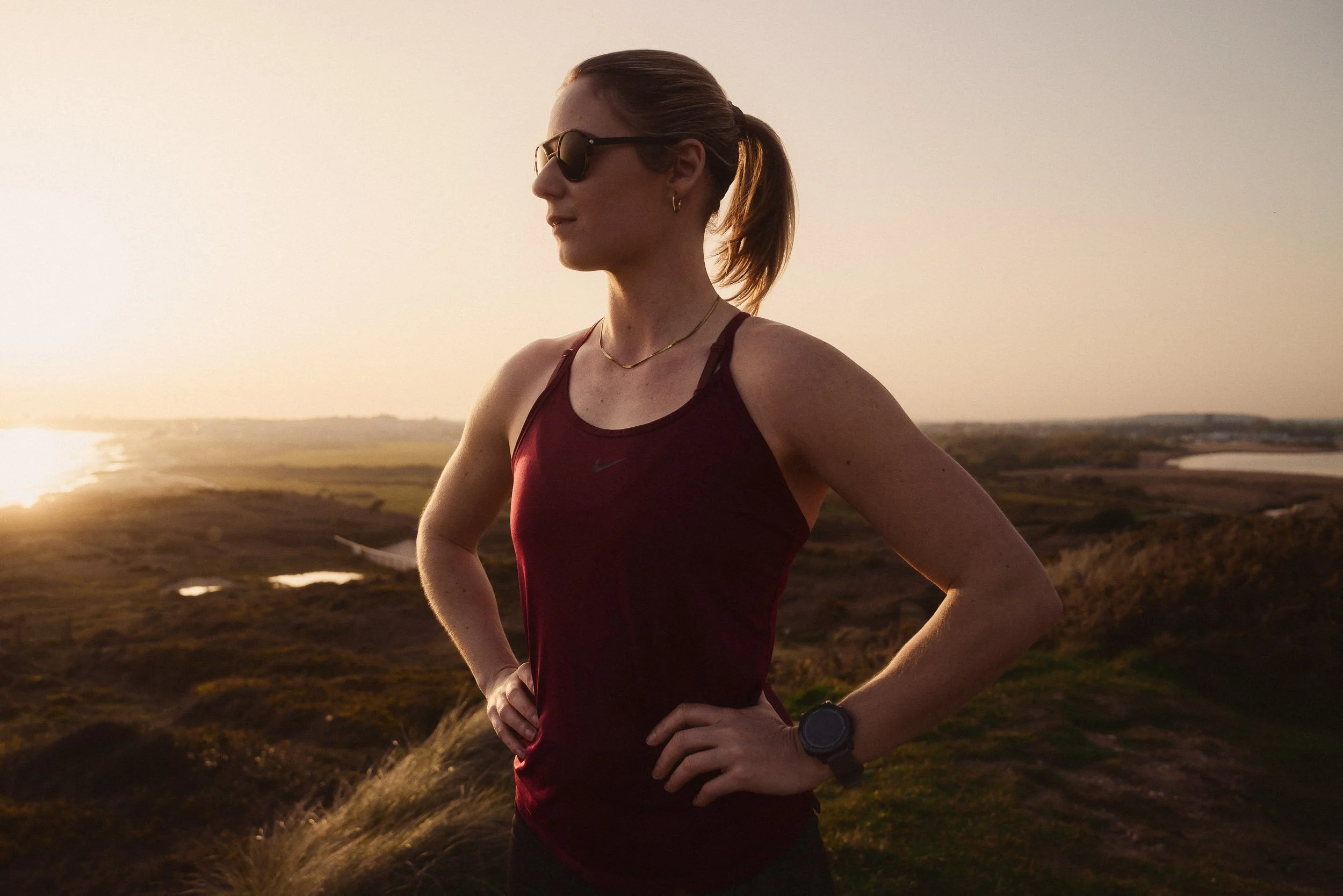 A portrait of a female athlete outdoors during sunset, wearing sunglasses, a maroon sports top, and a wristwatch, with a scenic landscape in the background.