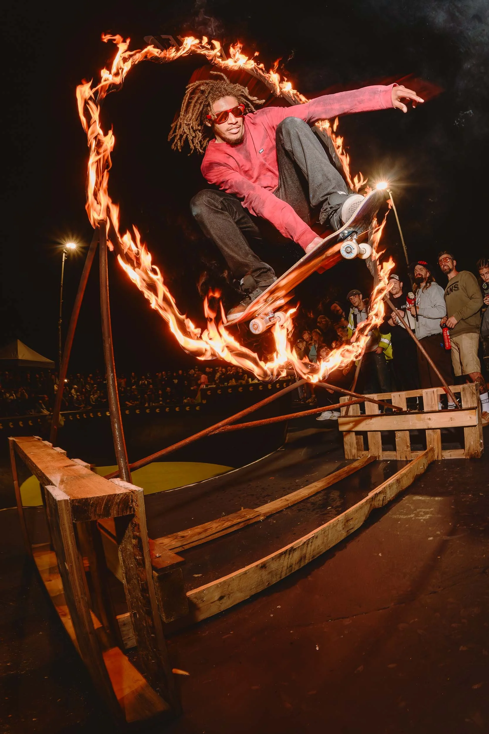 skateboarding performing skateboarding trick through a ring of fire at night, surrounded by onlookers.