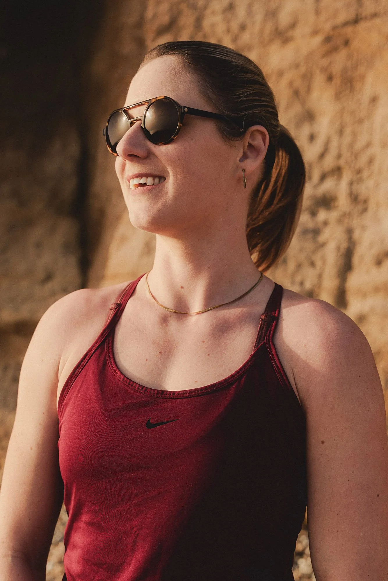 An athlete wearing sunglasses, a red Nike sleeveless athletic top, a gold necklace, and earrings, standing in front of a rock formation outdoors.