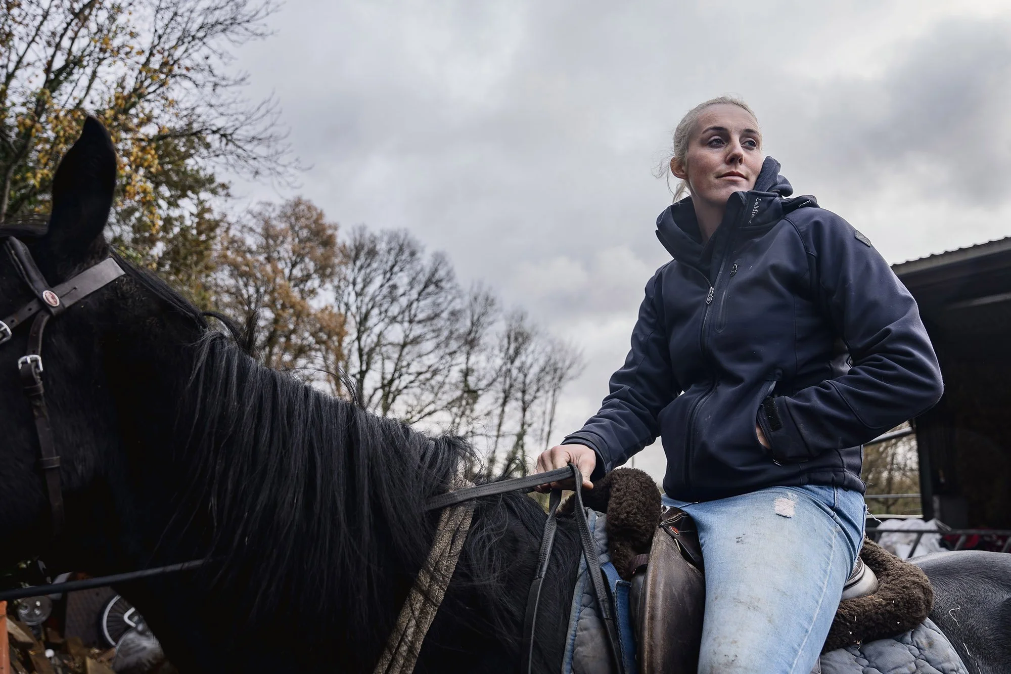 A woman riding a black horse outdoors on a cloudy day, wearing a navy jacket and blue jeans.