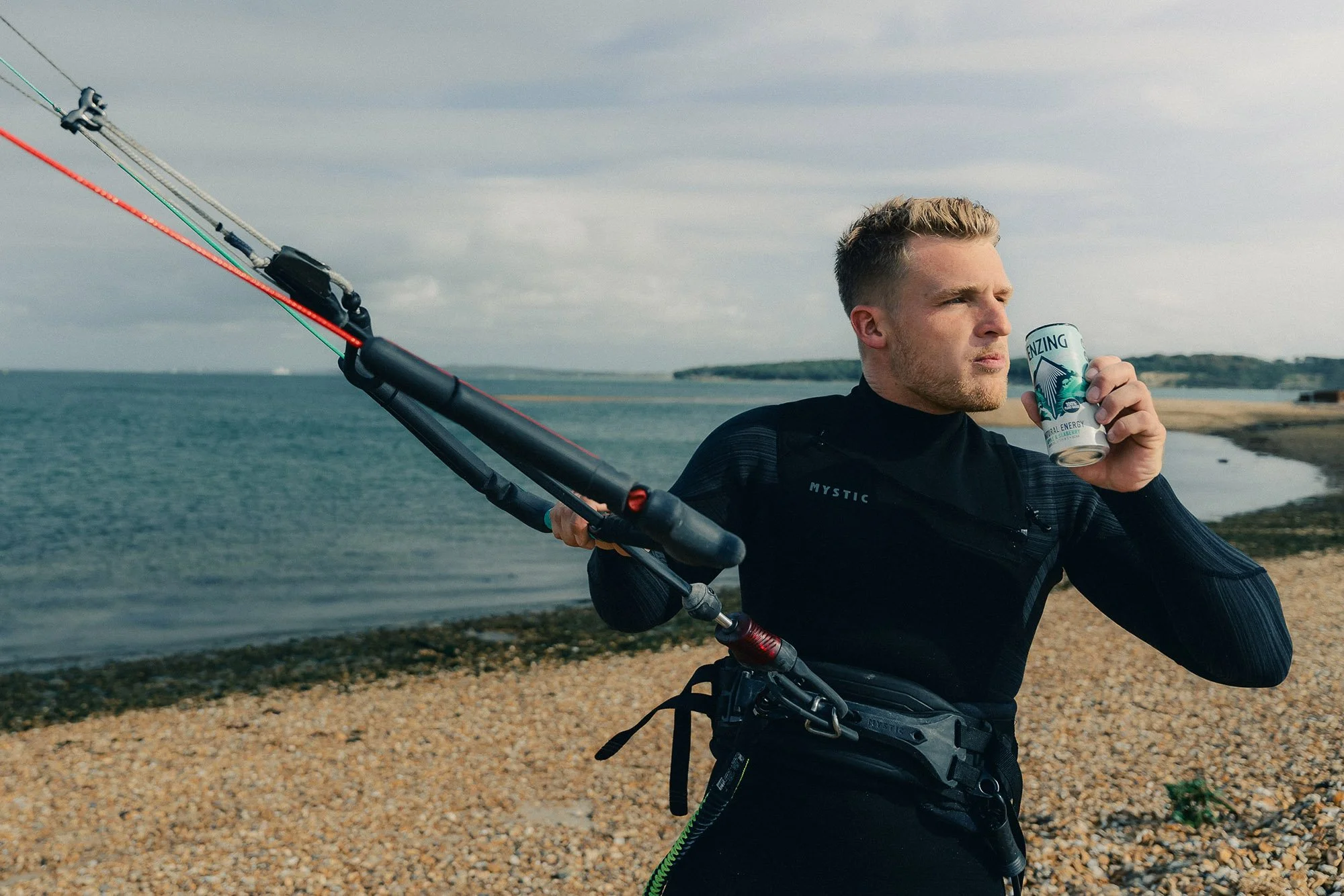A man in a black wetsuit standing on a rocky shoreline, holding a kite surfing control bar while drinking from a can, with the ocean and cloudy sky in the background.