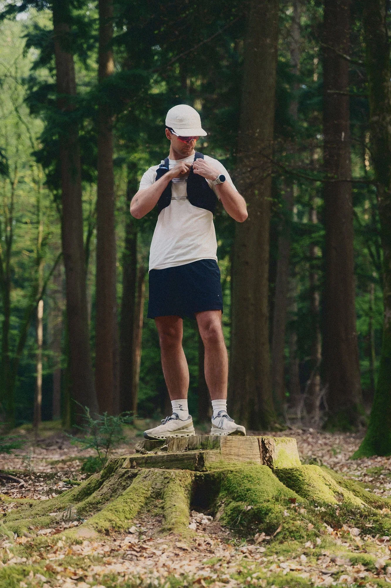 An athlete dressed in running gear, including a white cap, sunglasses, a light-coloured t-shirt, dark shorts, and running shoes, stands on a tree stump in a forest. He is adjusting his Kiprun running vest and is wearing a wristwatch.