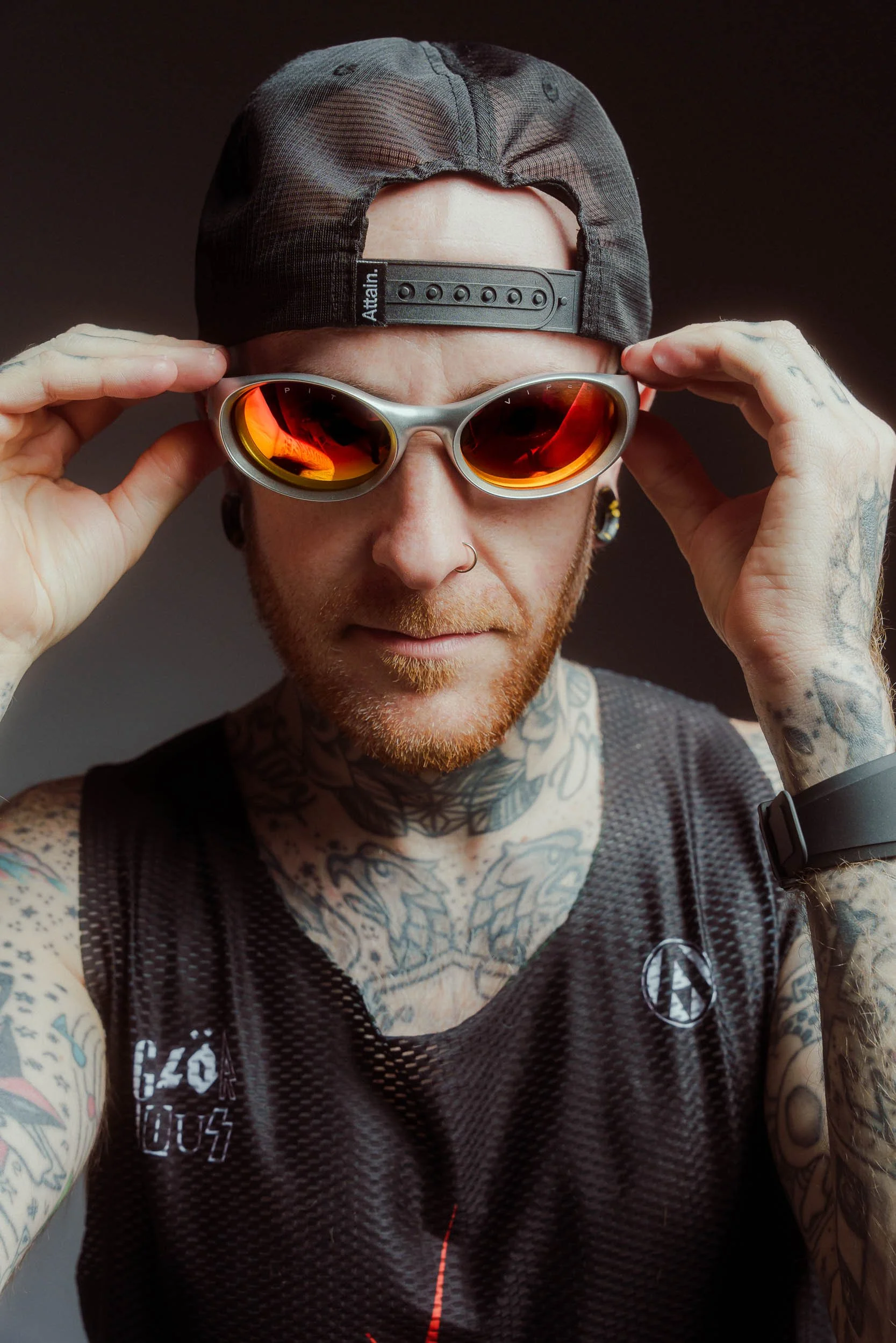 A male athlete wearing a black baseball cap, Pit Viper sunglasses, and a black mesh sleeveless shirt, adjusting his sunglasses with tattoos on his arms.