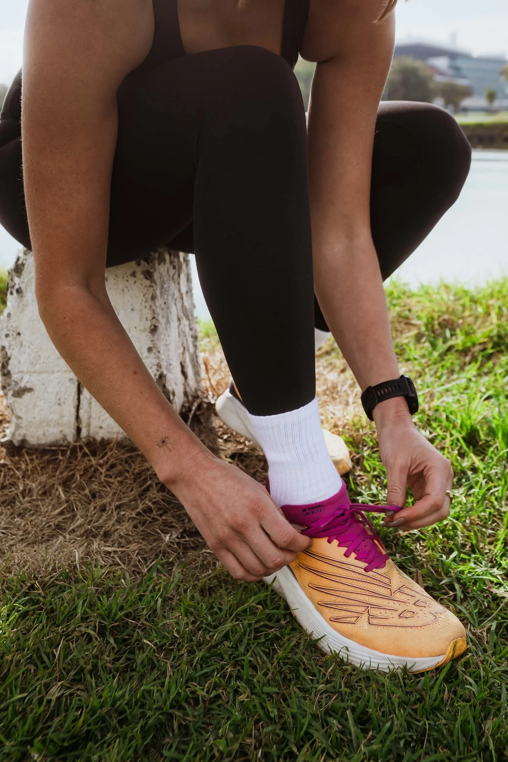 An athlete tying the laces of their orange and pink New Balance running shoes while wearing white socks, black leggings, and a black wristwatch, outdoors near a grassy area and a body of water.