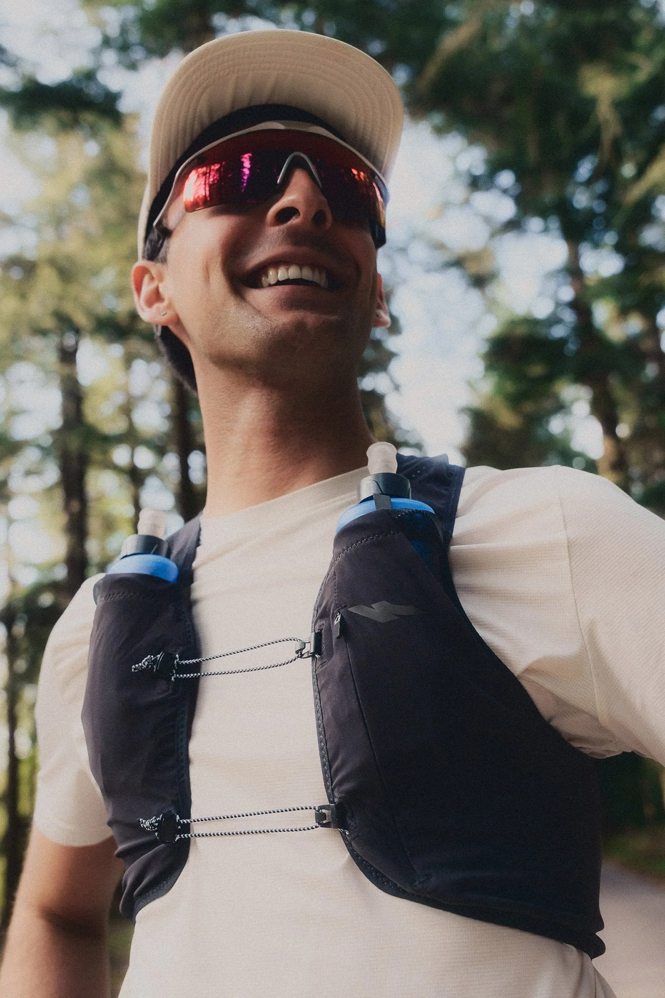 A runner smiling outdoors wearing sunglasses, a light-coloured hat, a white shirt, and a black kiprun running vest with soft flasks in pockets.