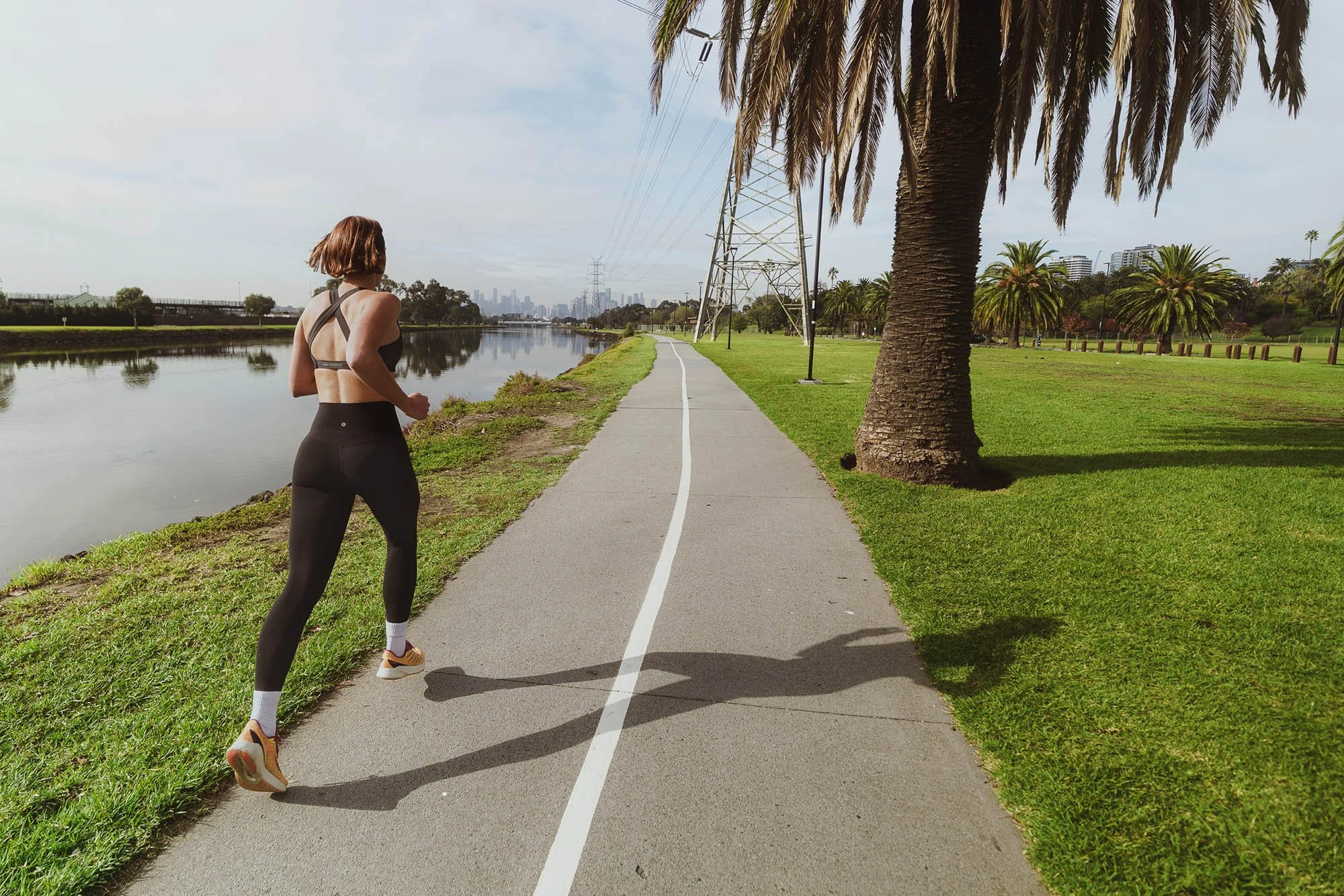 A woman jogging on a paved path beside a calm body of water in a park, with green grass, palm trees, and a city skyline in the background.