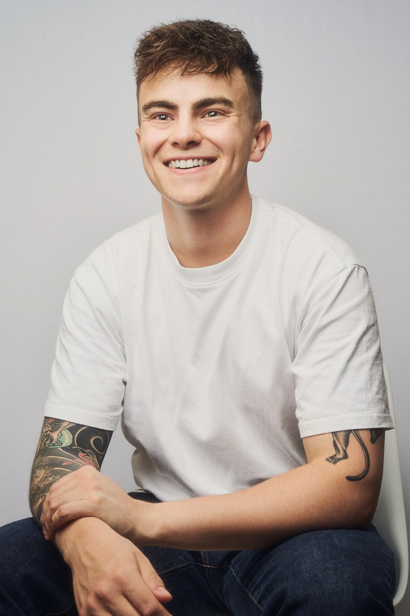 Photographer with short hair, wearing a white t-shirt, sitting with arms crossed and smiling, against a plain light gray background.