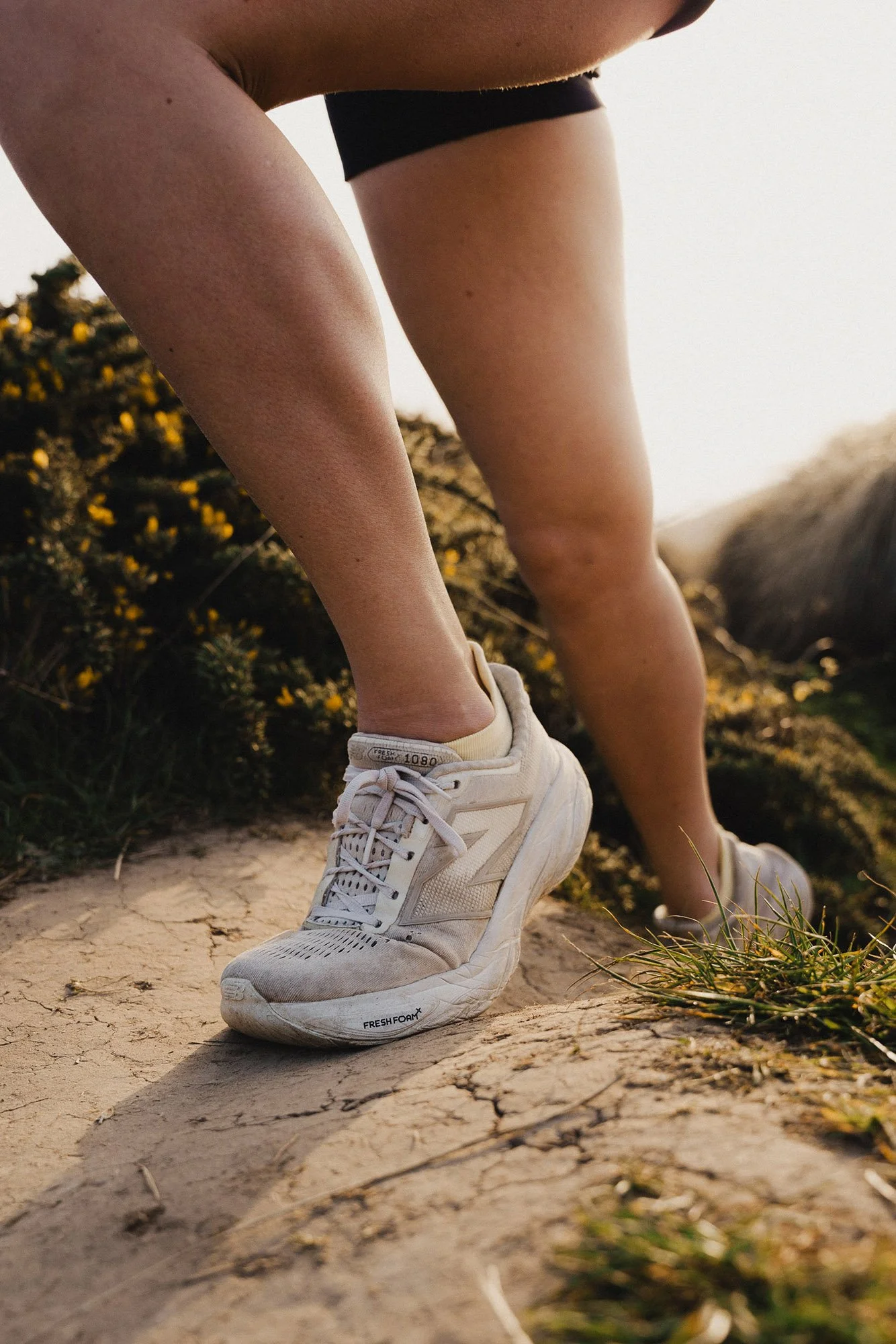 A person wearing white sneakers and black shorts, standing on a dirt trail with greenery and yellow flowers in the background.