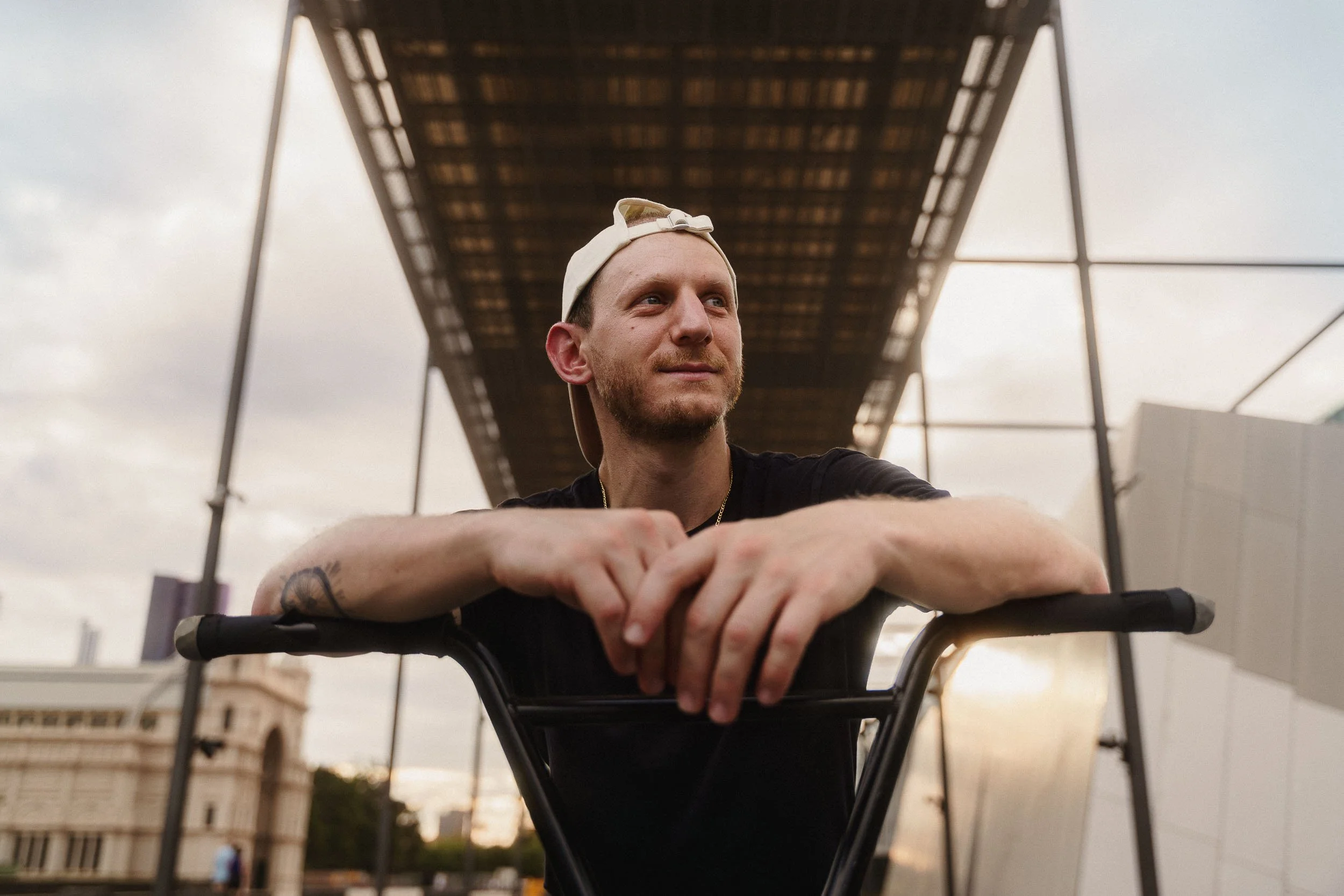 A BMX rider, wearing a beige cap backwards, is leaning on his BMX, looking into the distance. The background features modern architecture.