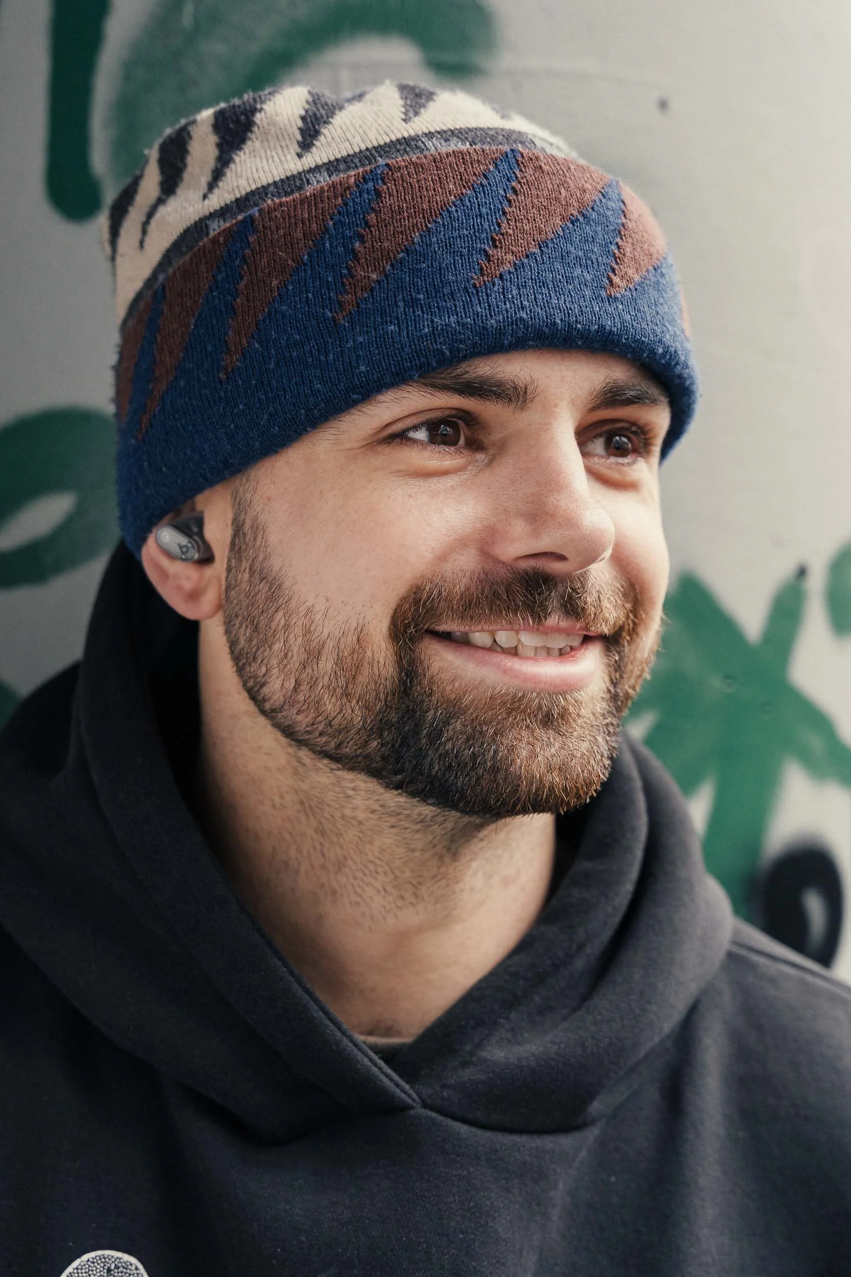 portrait of a smiling man wearing a colourful beanie and a black hoodie, with an earbud in his left ear, standing in front of a wall with graffiti on it.