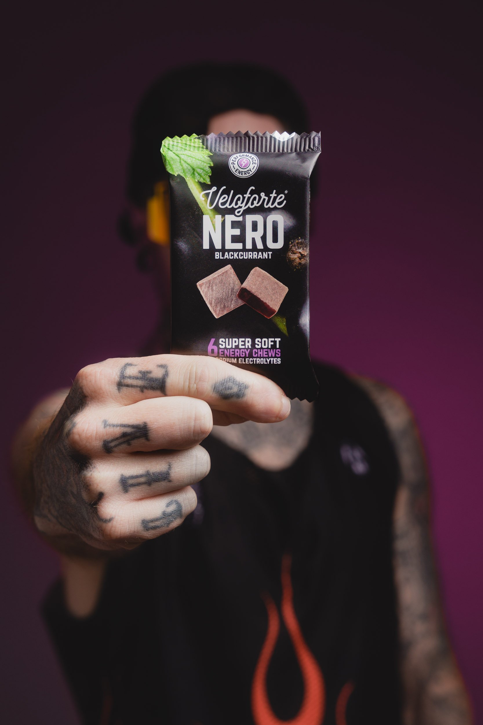 Person holding a black package of Veloforte Nero Blackcurrant chews with purple and brown accents, with tattoos on their hand and a blurred background.