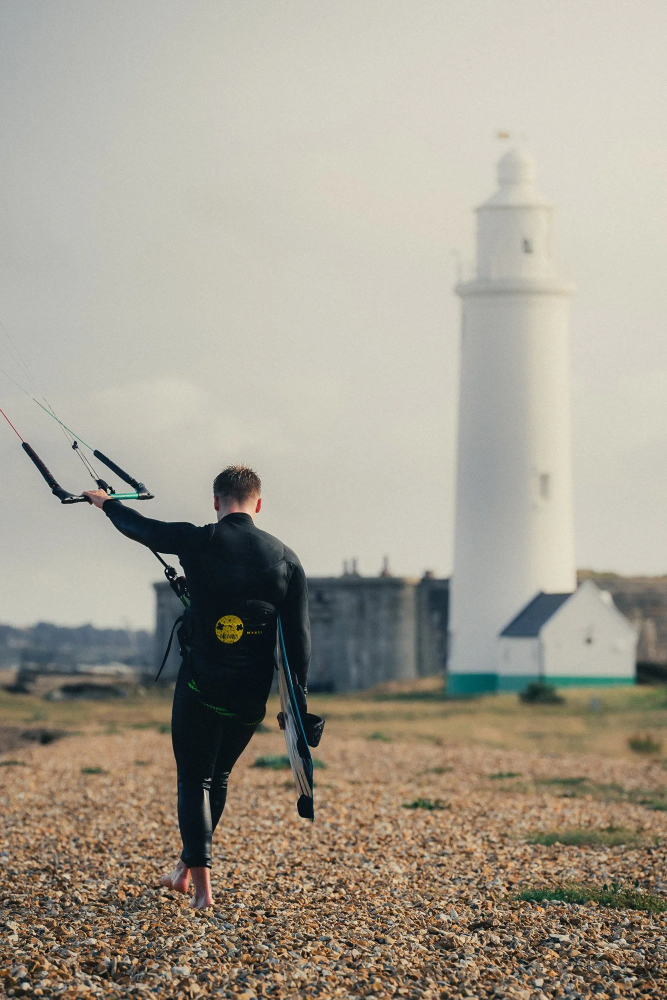 A kitesurfer walking barefoot on a pebble beach carrying a kiteboard with a lighthouse in the background.