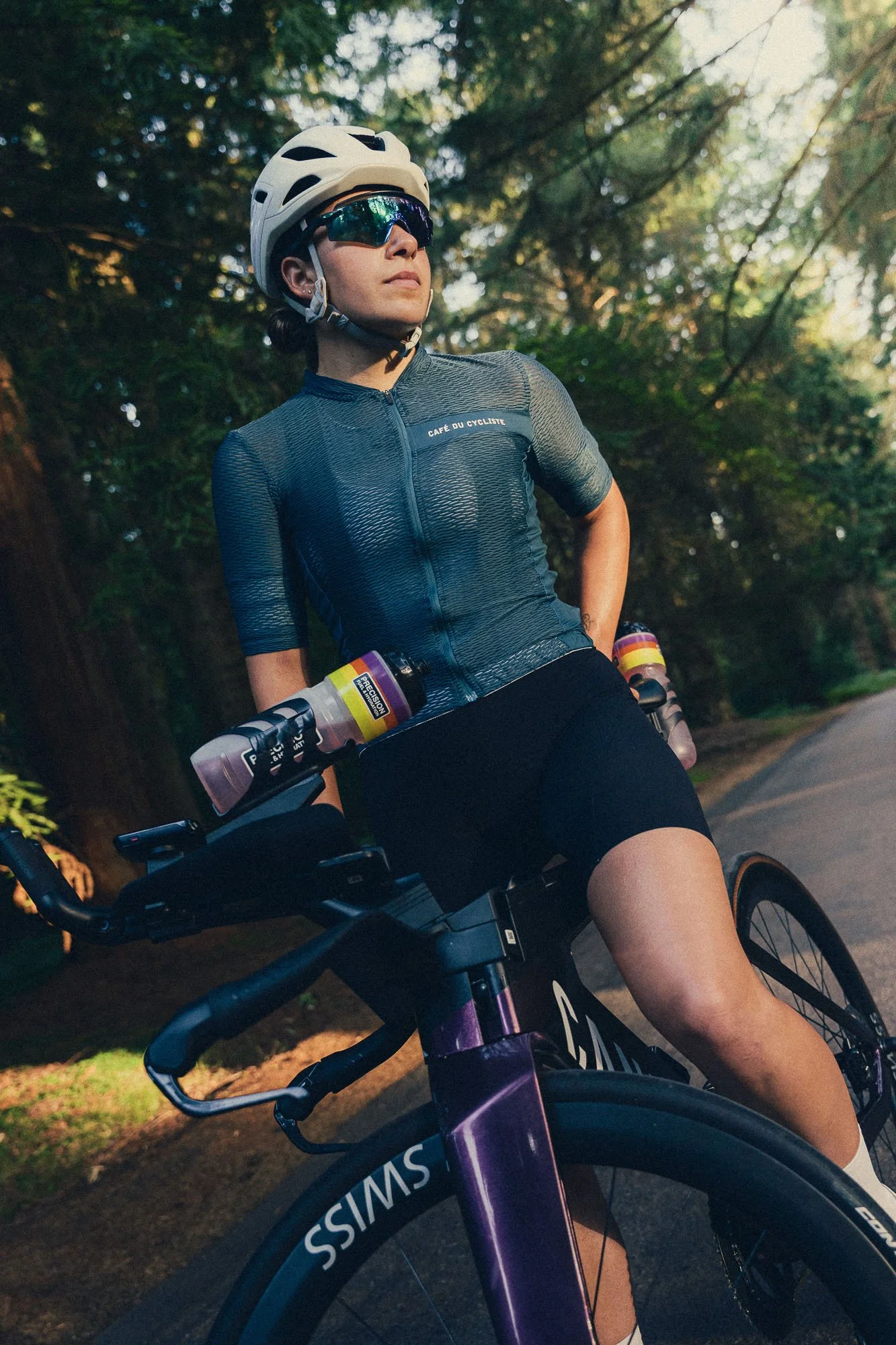 A woman wearing a white helmet and sunglasses, dressed in a dark cycling jersey, is sitting on a bicycle in a wooded area during daylight.