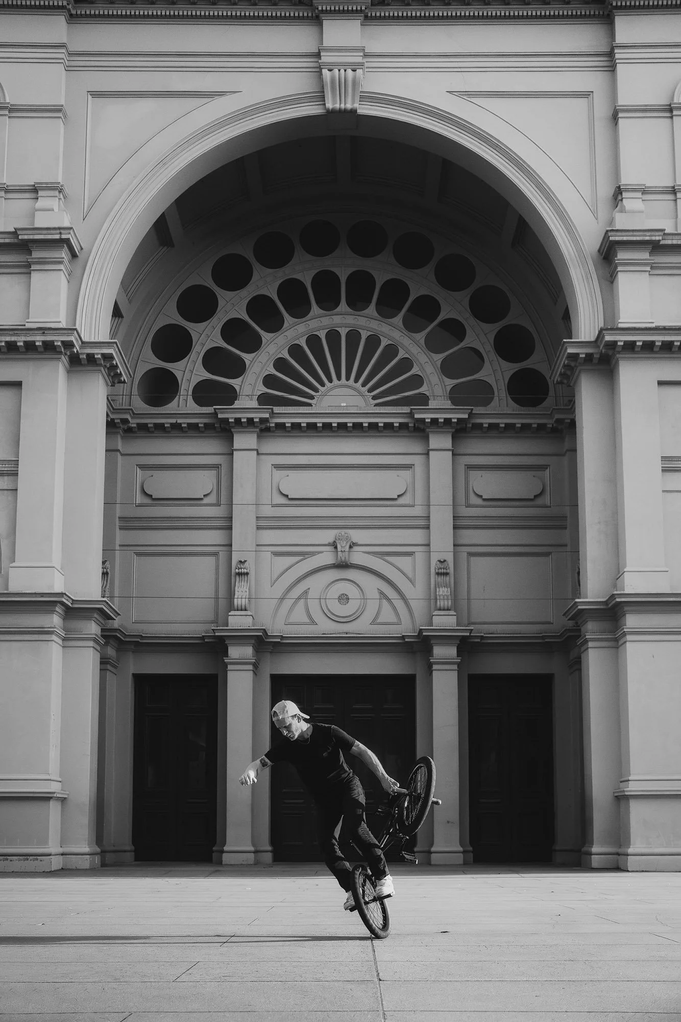 A person performing a BMX stunt in front of a historic building with large arched windows and ornate architectural details, in black and white.