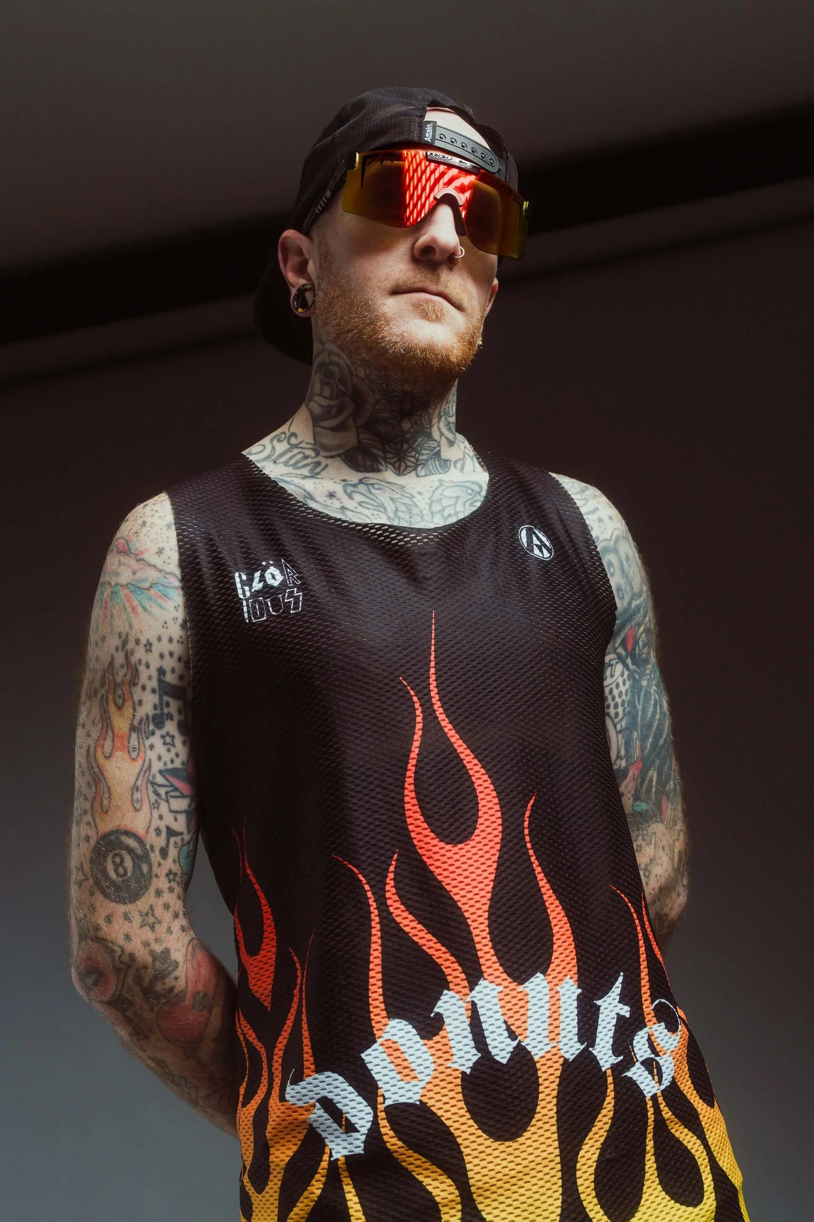 A male athlete with tattoos on his arms and neck, wearing Pit Viper sunglasses, a dark cap worn backwards, and a black athletic shirt with orange flames and bold white text.
