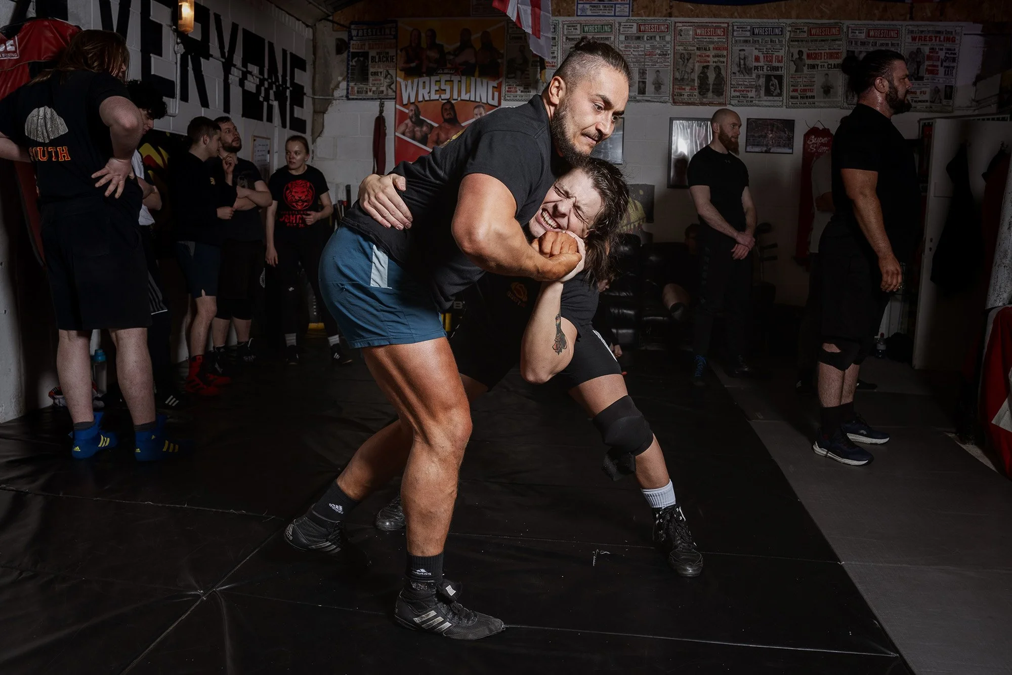 Two men wrestling on a mat in a gym with posters and banners related to wrestling on the walls. One man is in black athletic gear, and the other man is in black and gray clothing.