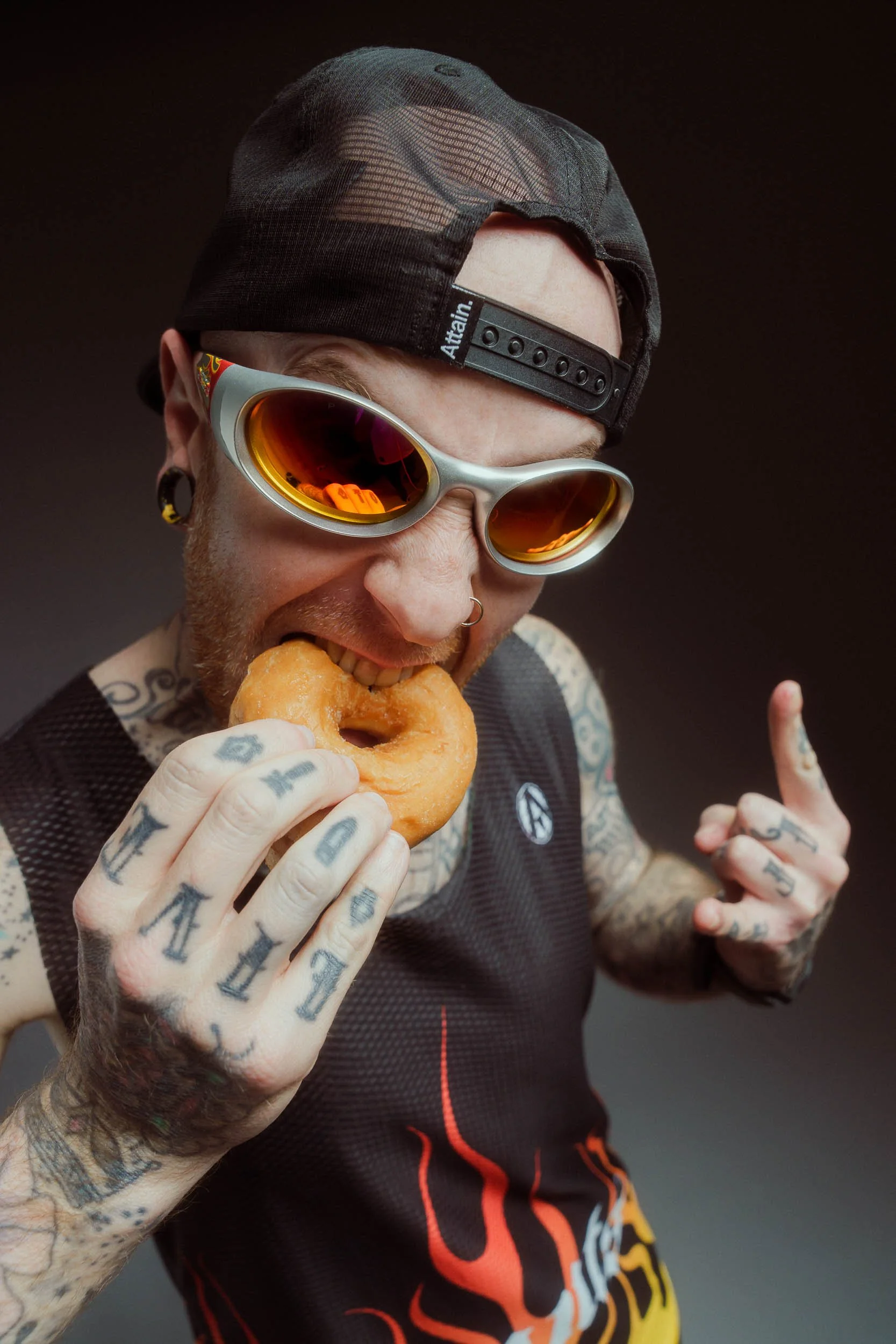 A person wearing a black mesh cap backward, orange mirrored sunglasses, and tattoos on their arms holding a donut with a bite taken out of it, and making rock hand signs with their fingers.