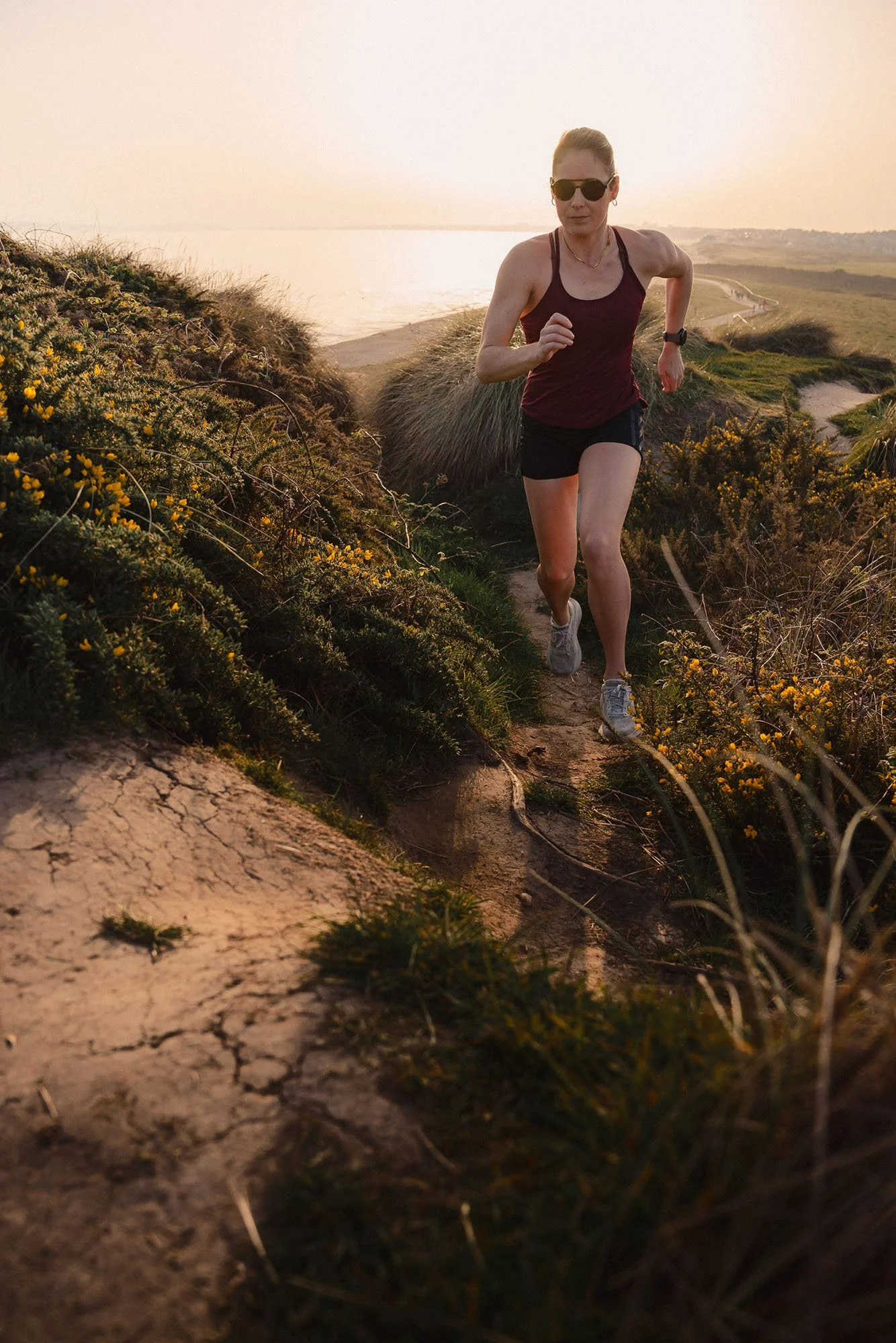 A female athlete running on a sandy trail near the sea, wearing sunglasses, a sleeveless shirt, and shorts, with the sun setting in the background.