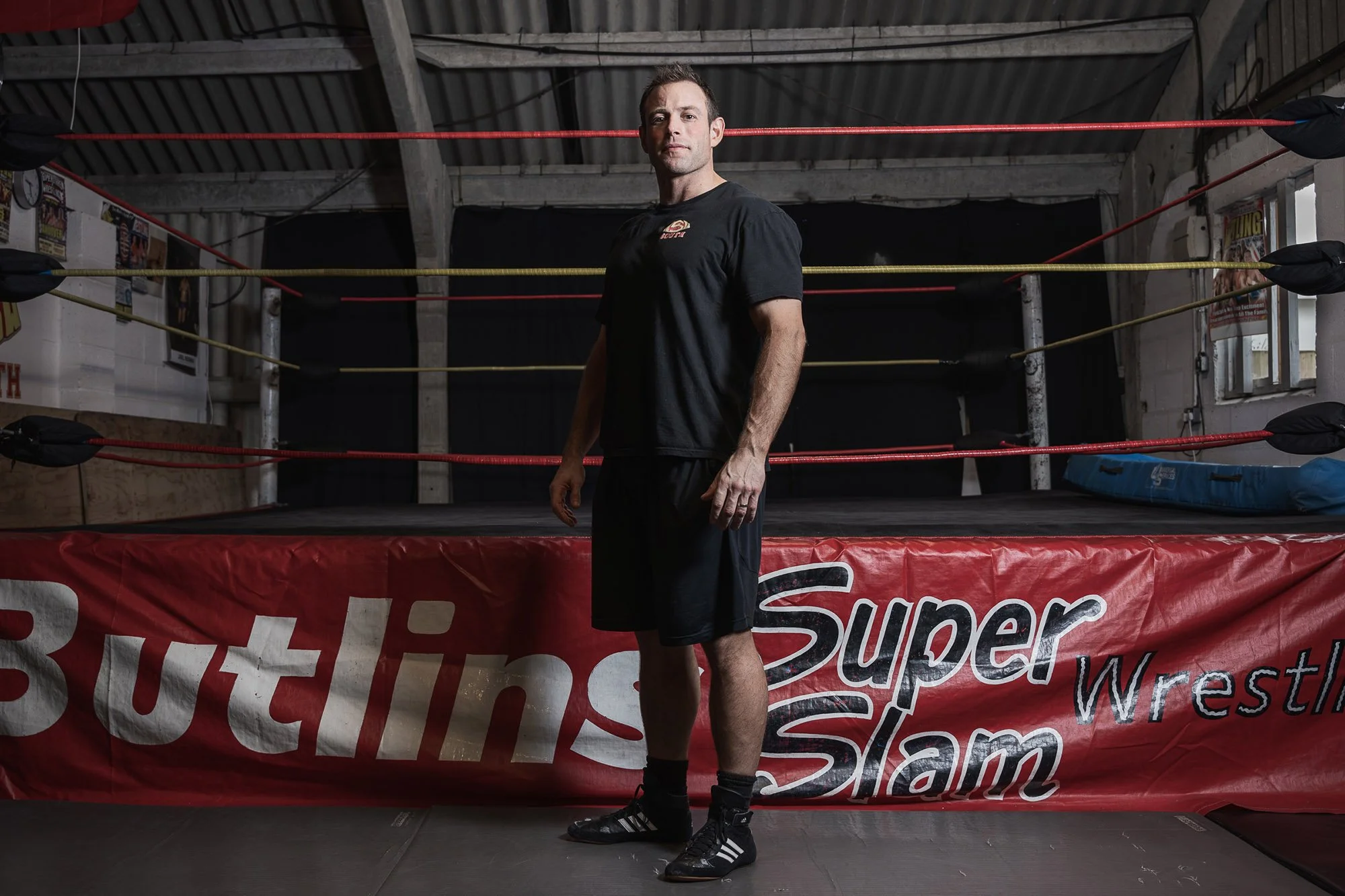 A man standing outside a wrestling ring with colourful ropes, in a gym with posters on the wall and a red banner on the ring