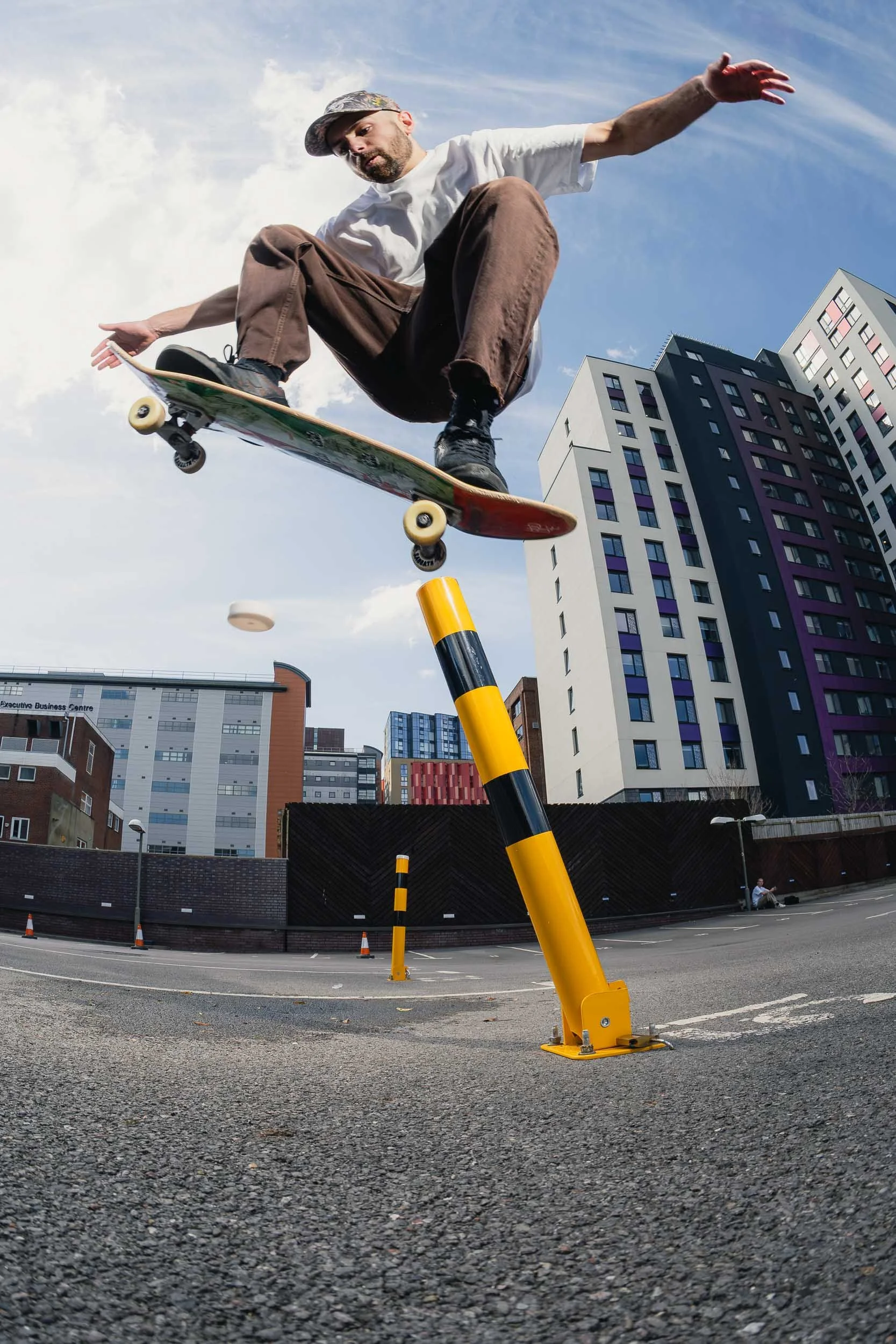 A skateboarder is performing a trick in mid-air over a parking lot and urban buildings, with a blue sky background and a yellow parking barrier.
