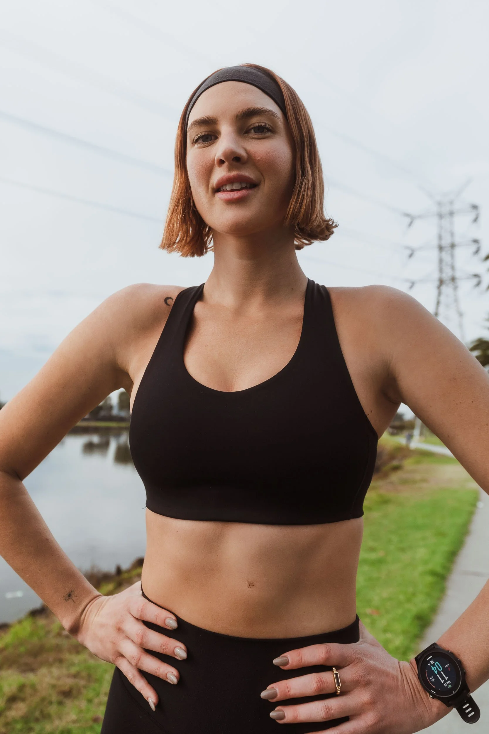 A female athlete in athletic wear standing outdoors with hands on her hips, near a pond, with power lines and trees in the background.
