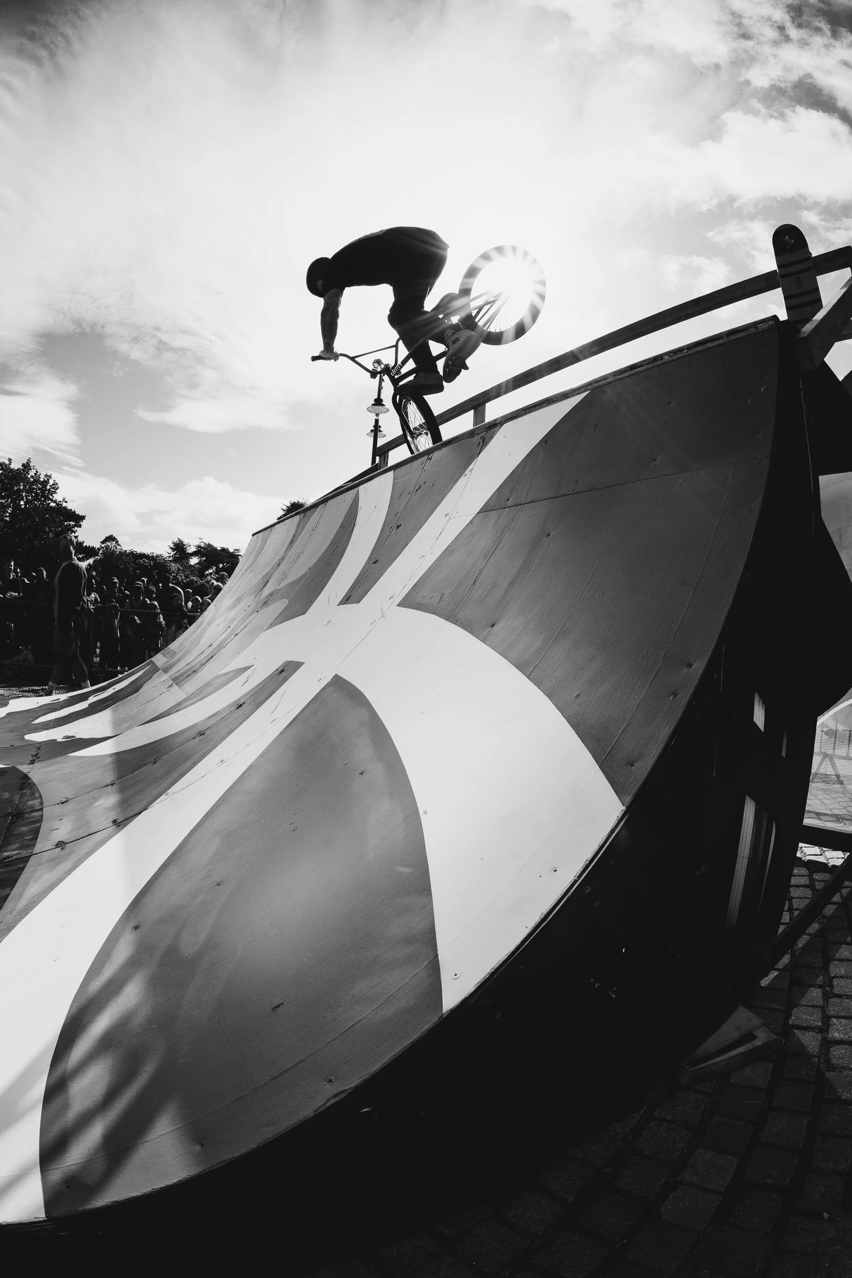 A person performing a trick on a BMX bike at a skatepark, with the sun shining through the bike's wheel, and a crowd of spectators in the background, captured in black and white.