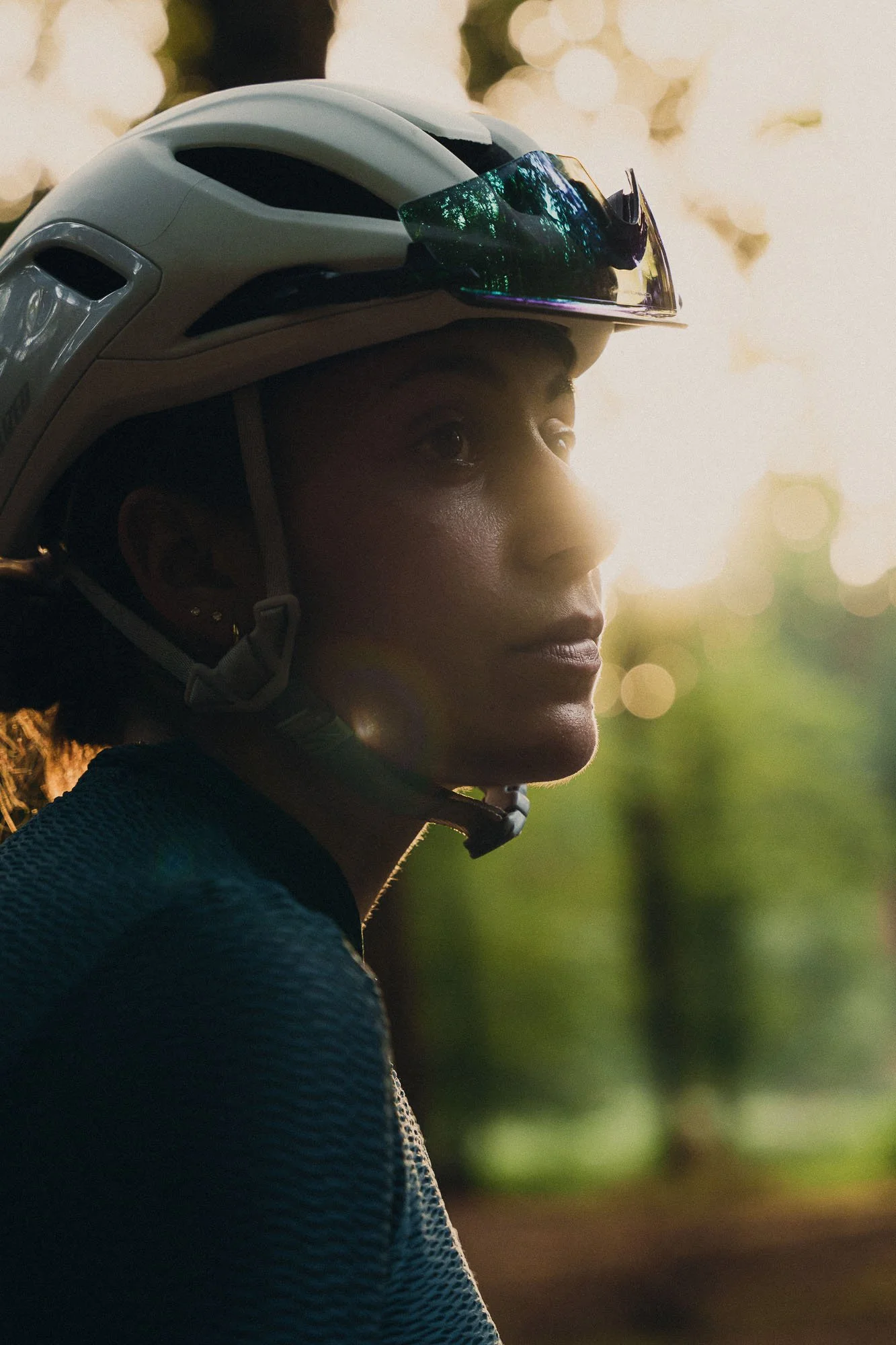 A woman wearing a white helmet and sunglasses on top, looking to the right, against a background of blurred greenery and soft sunlight.