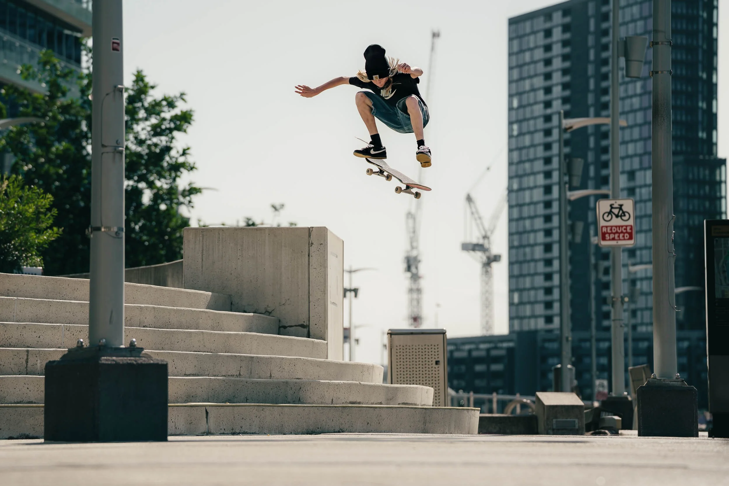 A person performing a skateboarding trick jumps over concrete stairs in an urban area with tall buildings and construction cranes in the background.