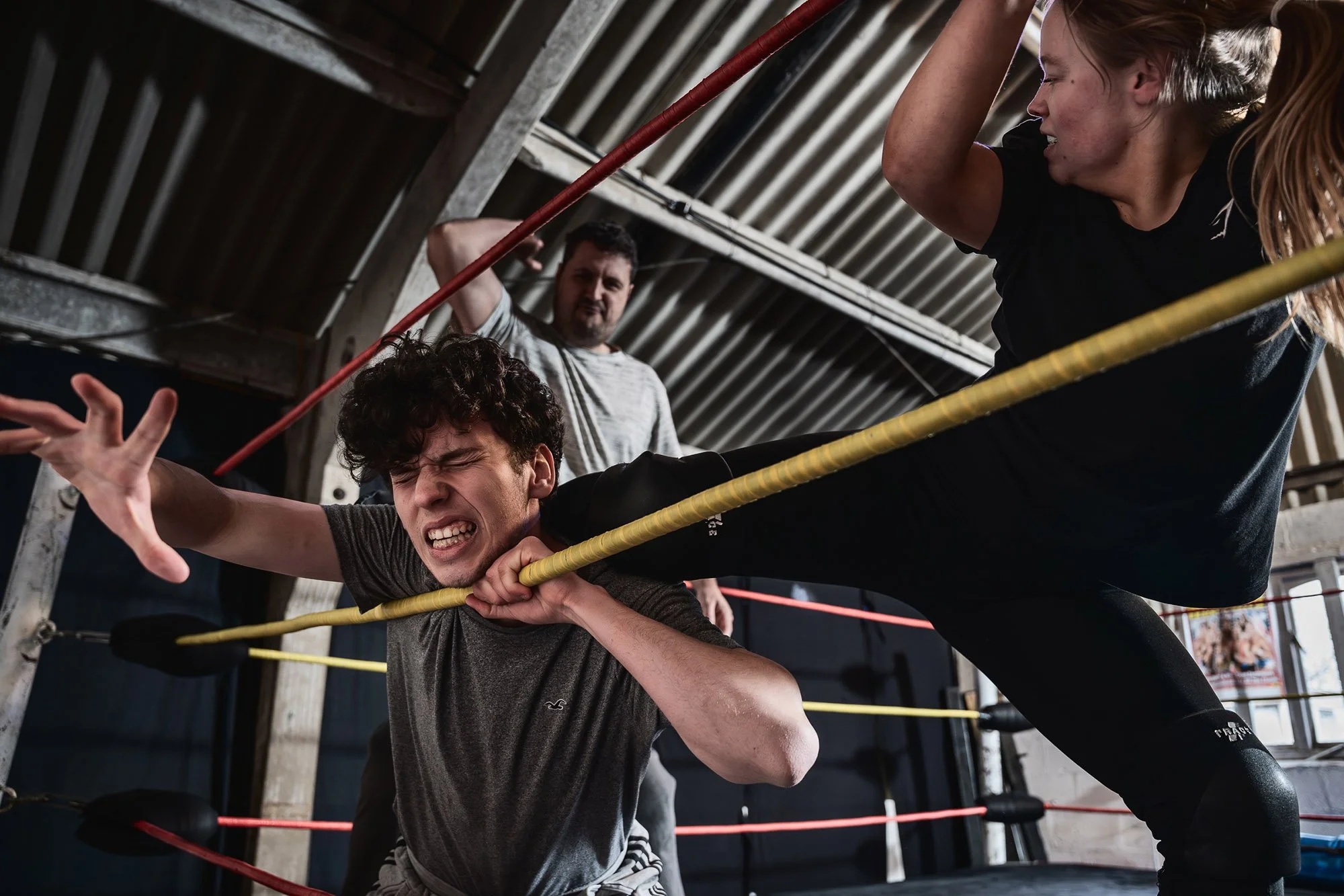 A female wrestler with a ponytail and a man with curly hair and a black shirt in a wrestling ring. The woman is leaning over the yellow ropes and kneeling on the man's neck, while the man is grimacing from the contact and holding onto the ropes.