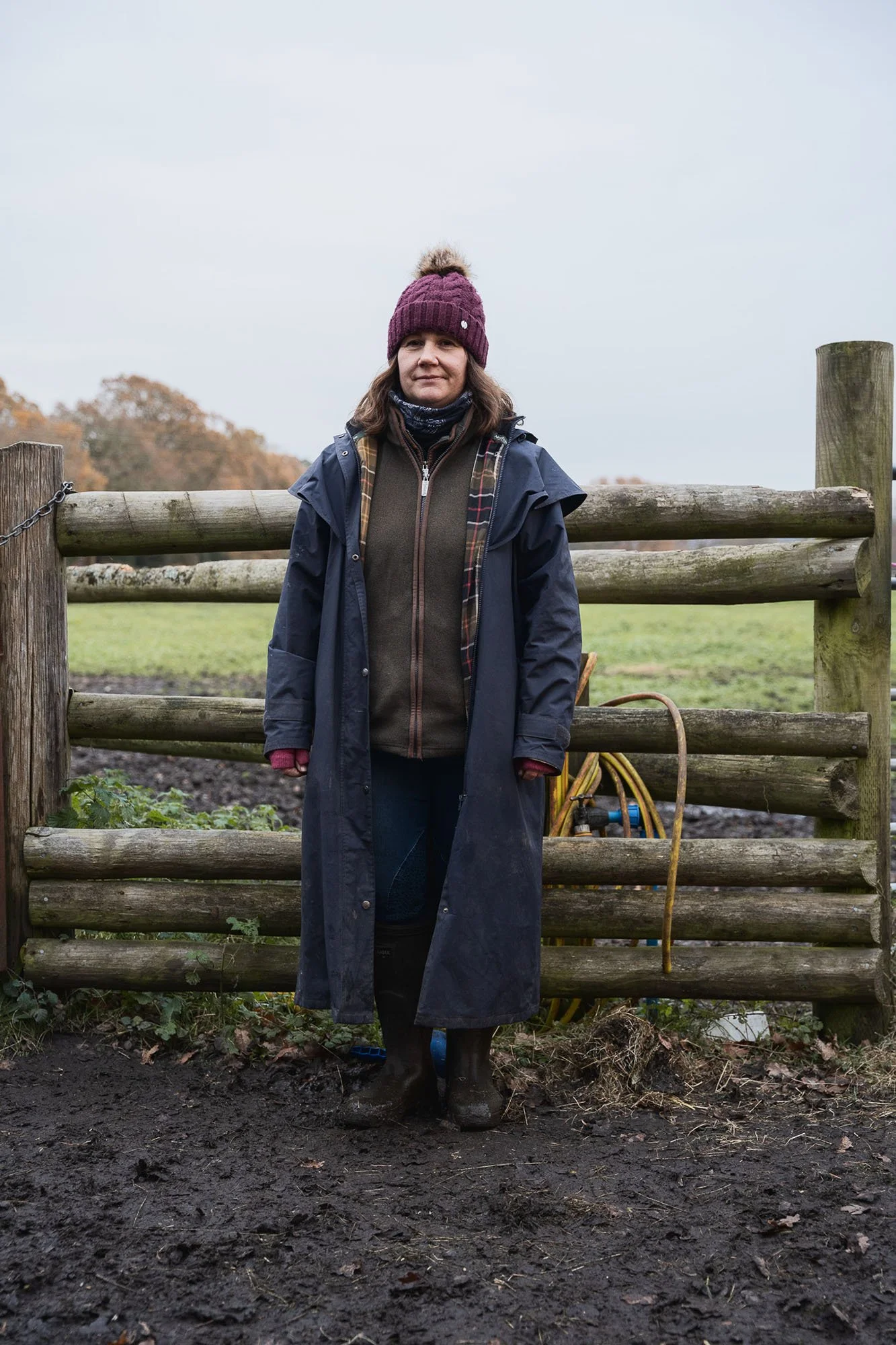 A woman standing outdoors behind a wooden fence, wearing a purple knit hat, a dark raincoat, and rubber boots, with muddy ground and overcast sky in the background.