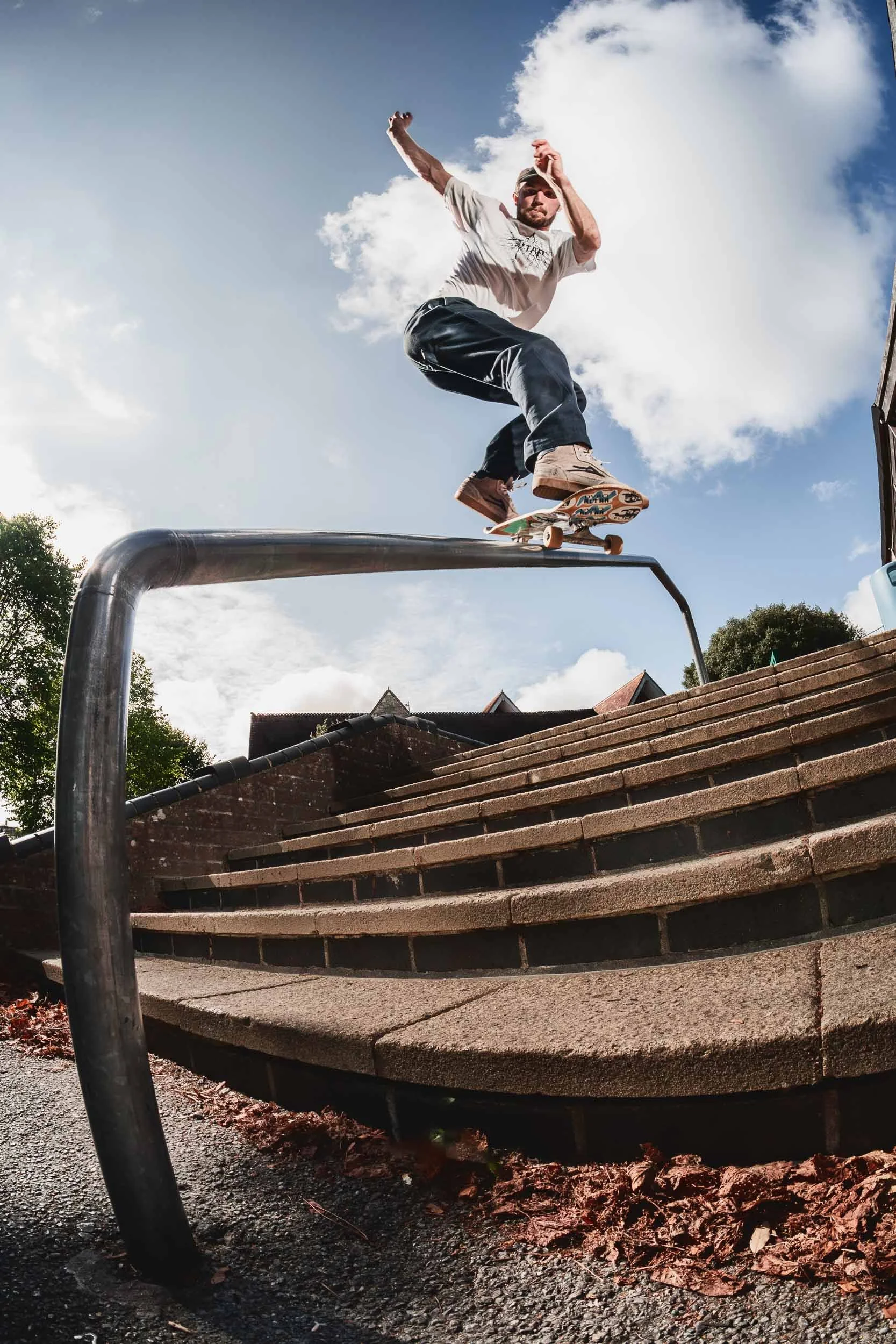 A person skateboarding on a ledge outdoors during the day, with stairs and trees in the background, under a partly cloudy sky.
