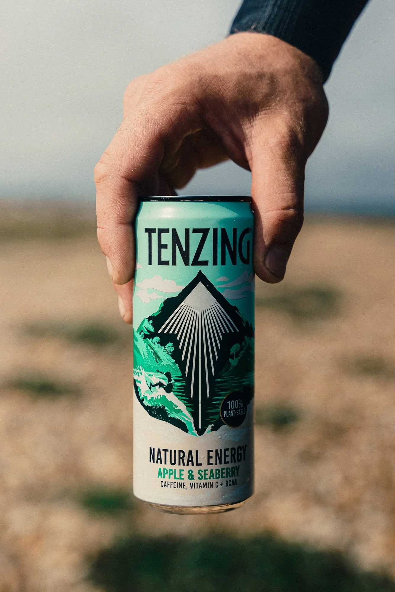 A hand holding a can of Tenzing natural energy drink, outdoors with a beach and sky in the background.