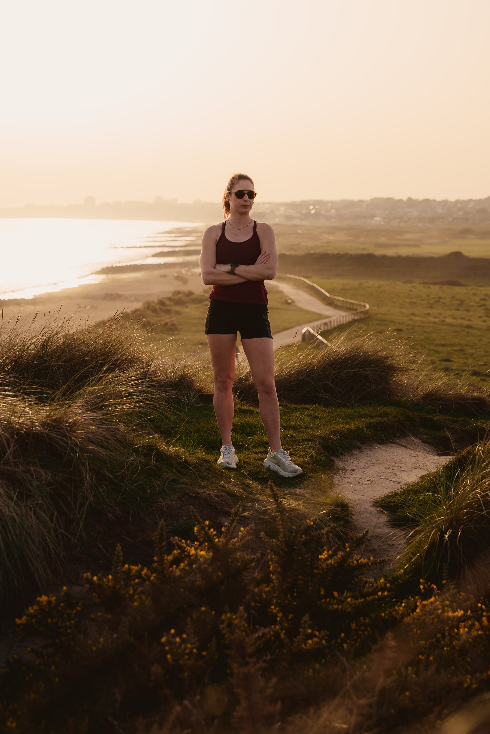 A female athlete with sunglasses, wearing a tank top, shorts, and running shoes, standing on a grassy hill with arms crossed, during sunset, overlooking a coastal landscape with a winding pathway and distant water.